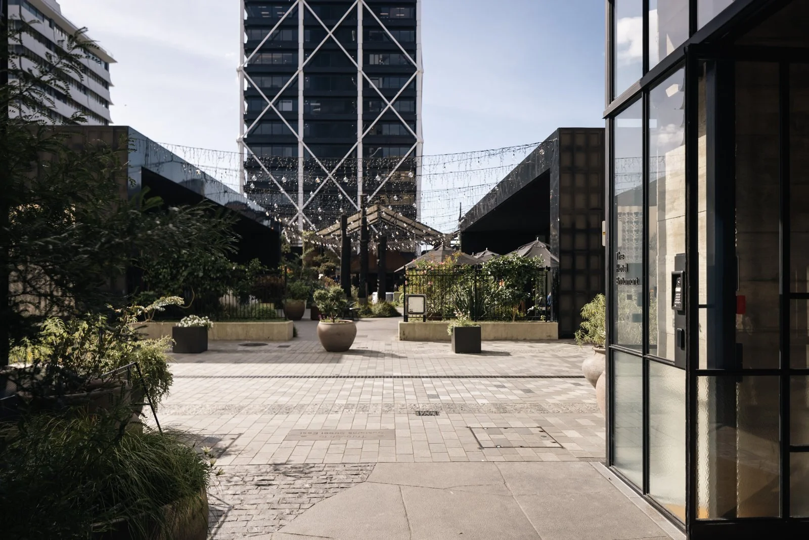 A modern outdoor urban patio area with potted plants and string lights, surrounded by tall buildings, including a high-rise with a white lattice structure, and adjacent to an entrance with glass doors. Shadows and a partly cloudy sky are visible.