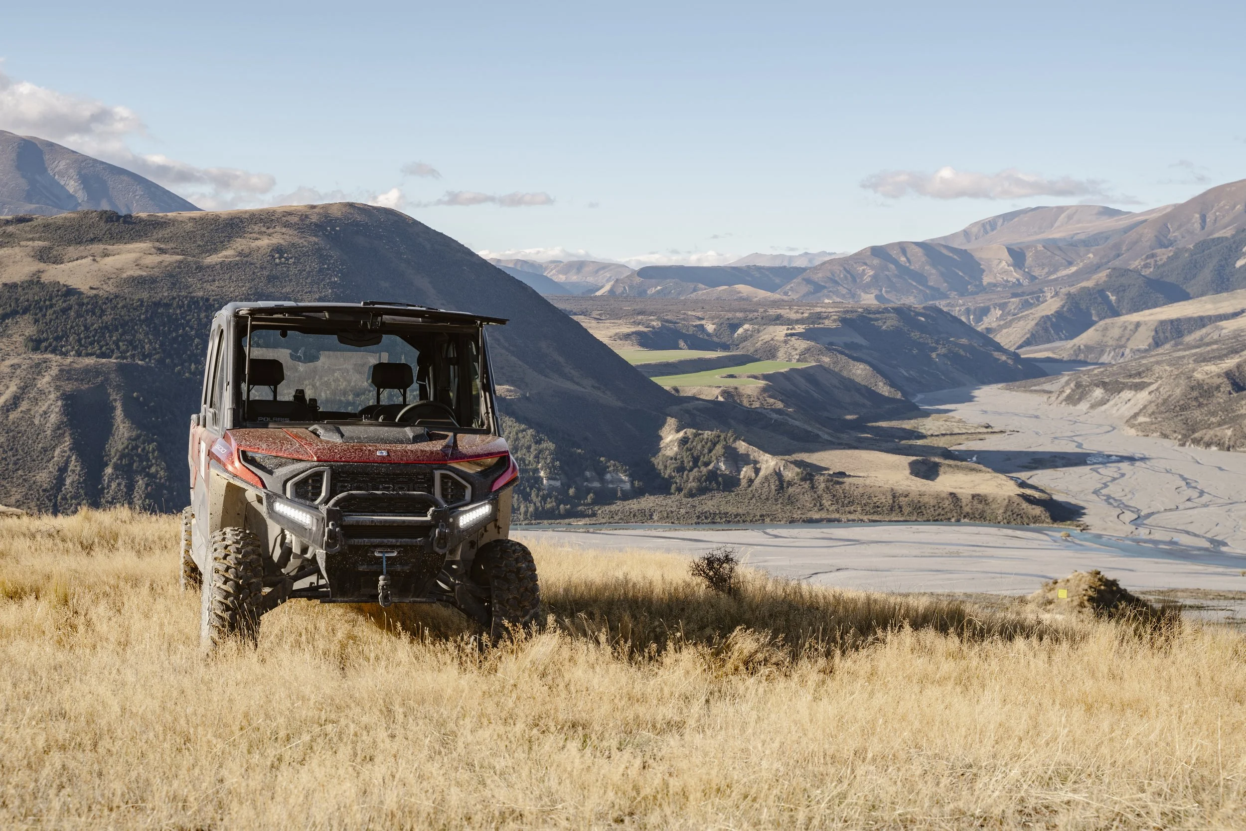 An off-road utility vehicle parked on a grassy hill overlooking a deep valley with a winding river, surrounded by mountain ranges under a clear blue sky with scattered clouds.
