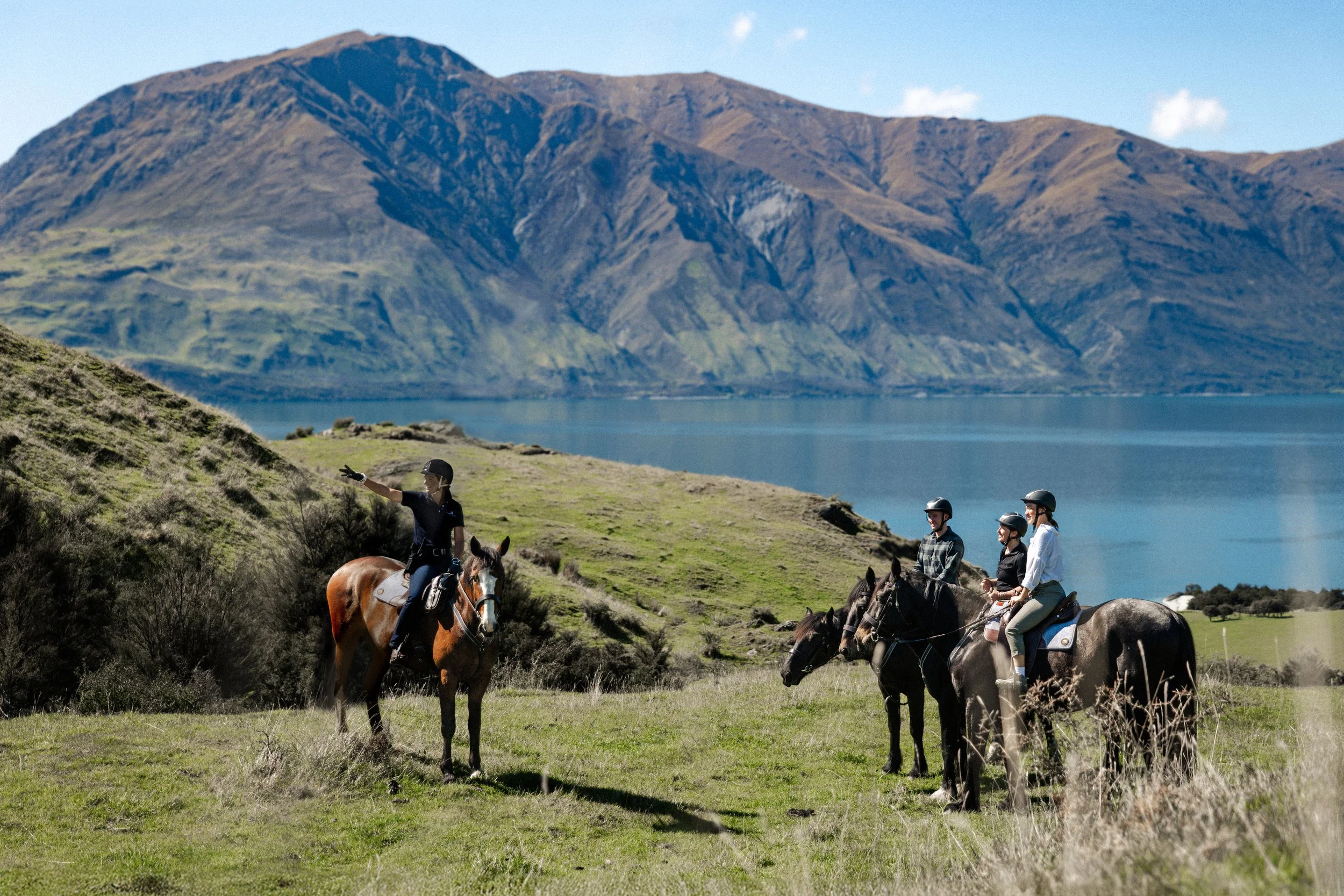 A group of children riding horses on green grass with mountains in the background, and a lake nearby.
