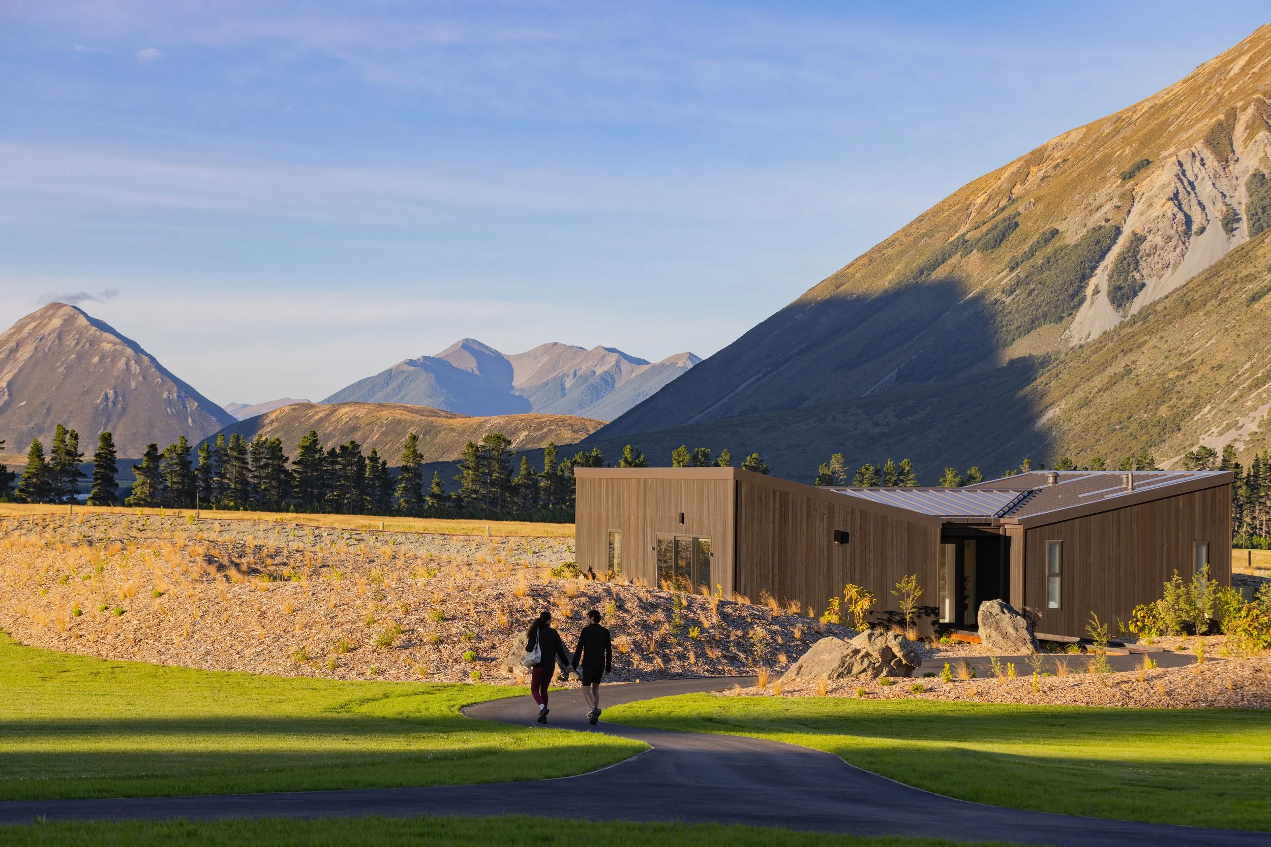 People walking on a paved path in front of modern houses with a backdrop of mountains under a blue sky.