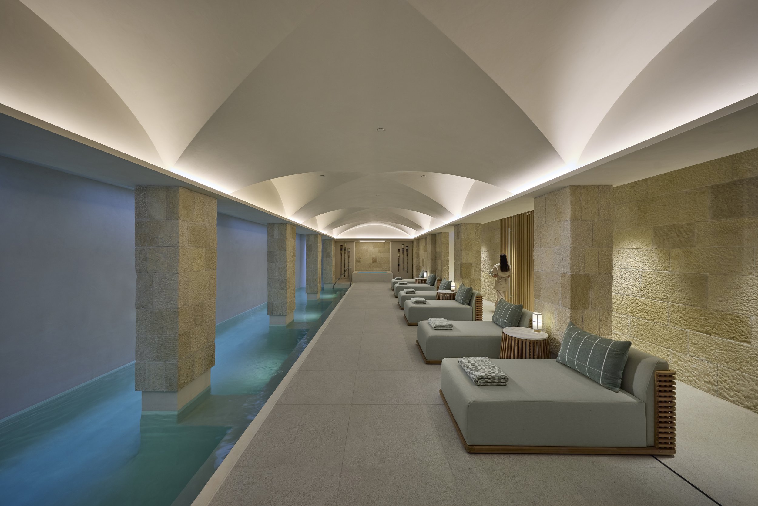An indoor pool area with a row of lounge chairs along the right side, a woman in a robe standing near the chairs reading, stone walls, arched ceiling with soft lighting, and a calm swimming pool.