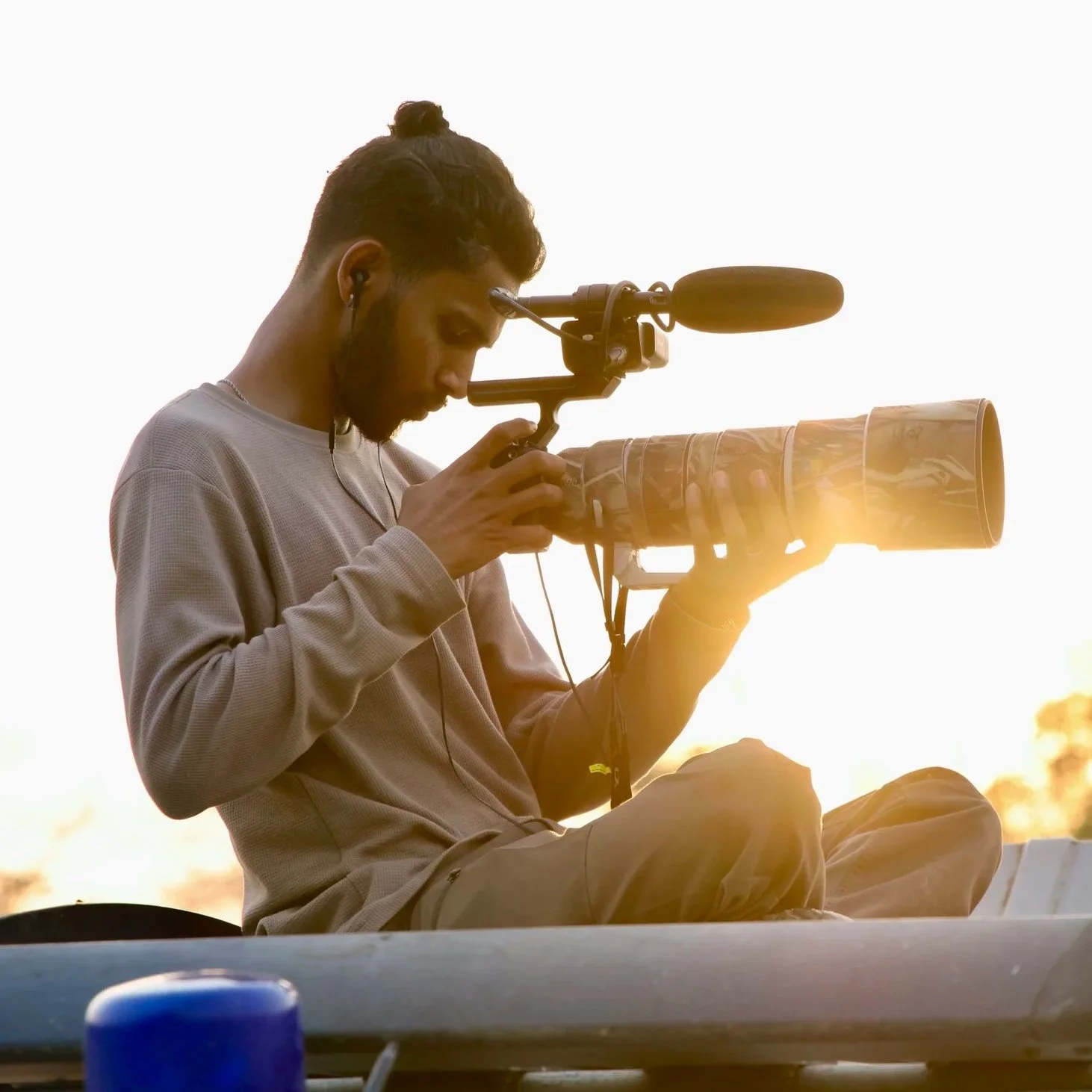A man sitting outdoors at sunset, looking through a large camera with a long telephoto lens, wearing earphones and casual clothing.