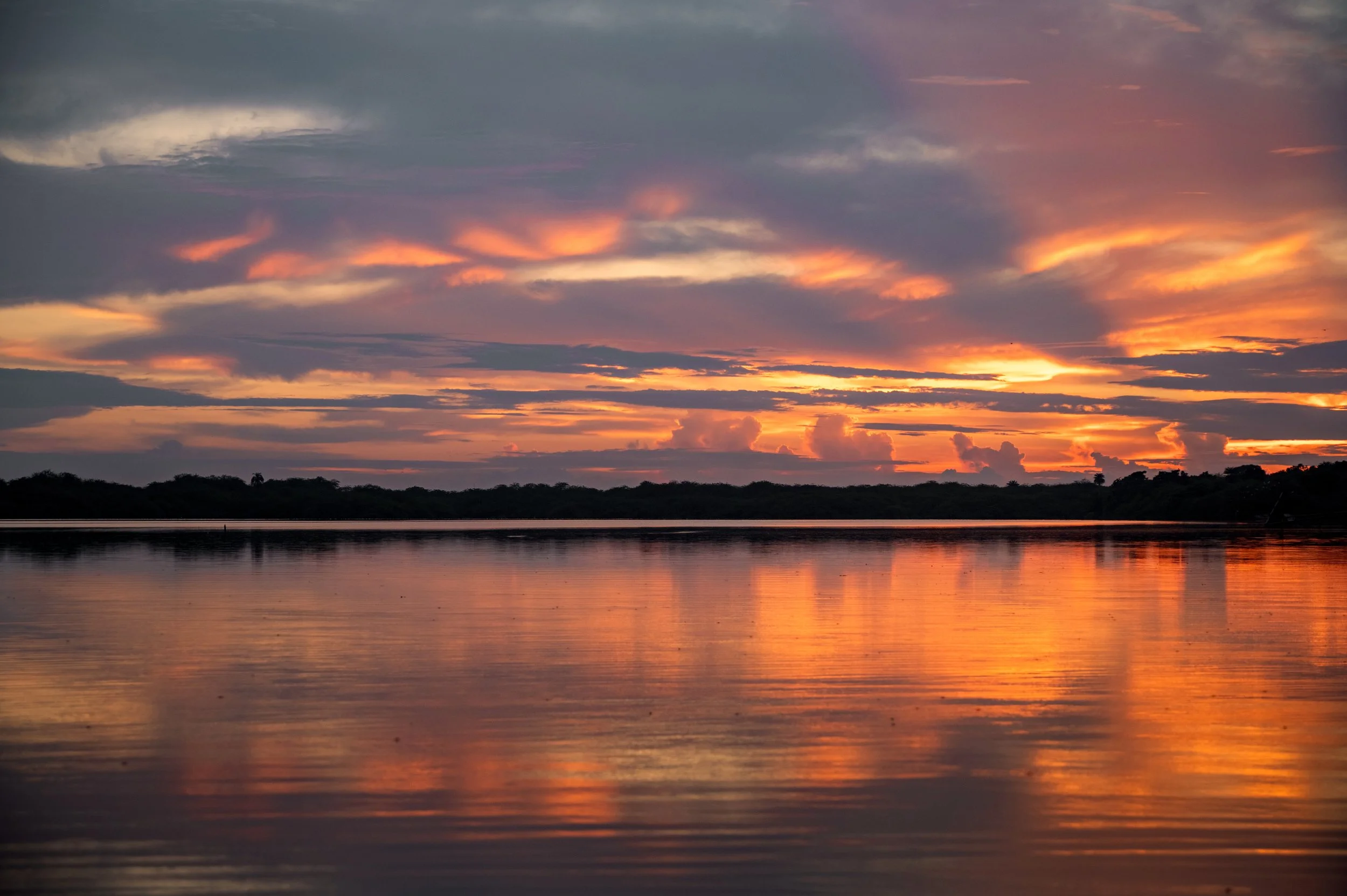 A serene sunset over a calm body of water, with colorful clouds in the sky reflecting on the water's surface and a silhouette of trees in the distance.