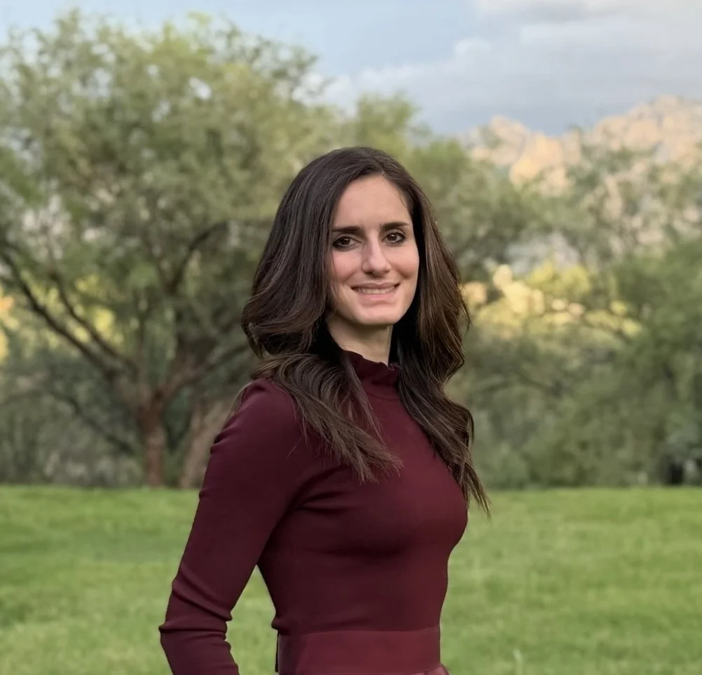 A woman with long brown hair and a maroon long-sleeve top stands outdoors in a park with green grass, trees, and distant mountains under a partly cloudy sky.