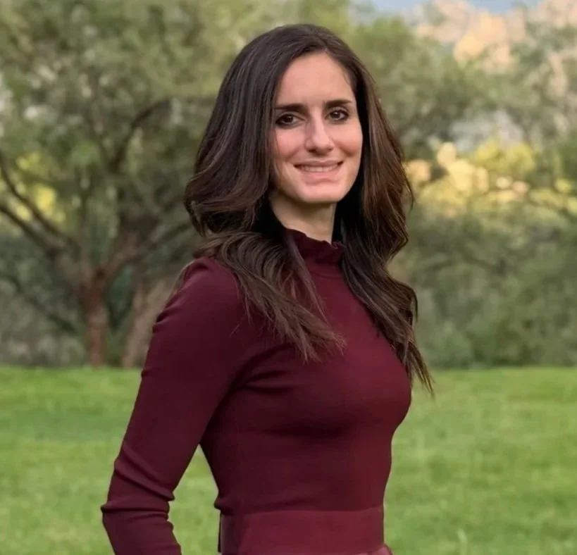 A woman with long dark hair smiling outdoors, wearing a maroon long-sleeve shirt with green trees and grass in the background.