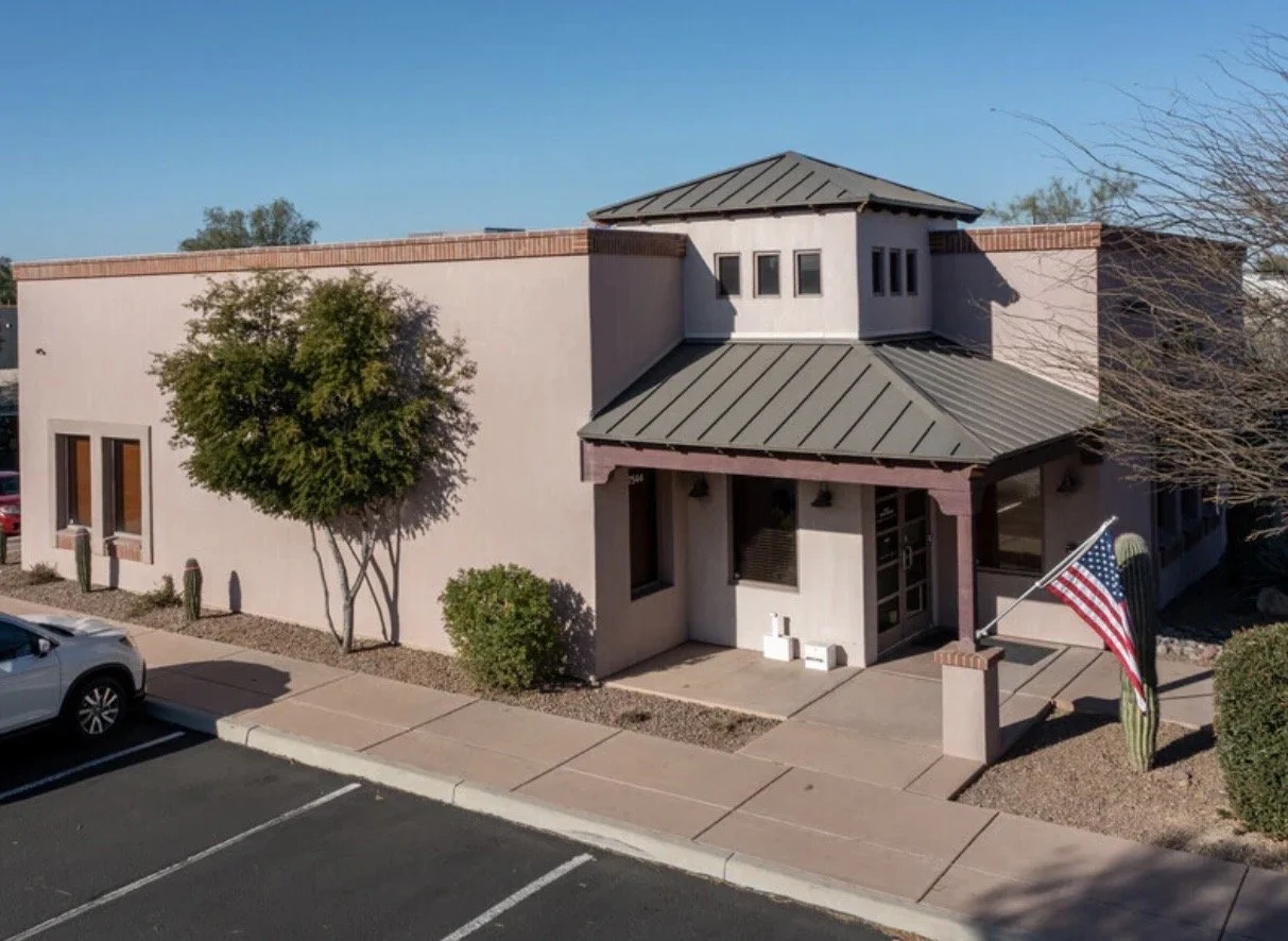 A modern building with a gray metal roof, a small covered entrance, a landscape with trees, cacti, and an American flag in front.
