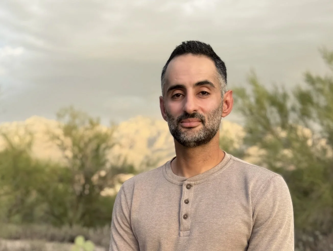 A man with short dark hair, a beard, and mustache standing outdoors during daytime with cloudy sky and green trees in the background, wearing a beige long-sleeve shirt with buttons near the collar.
