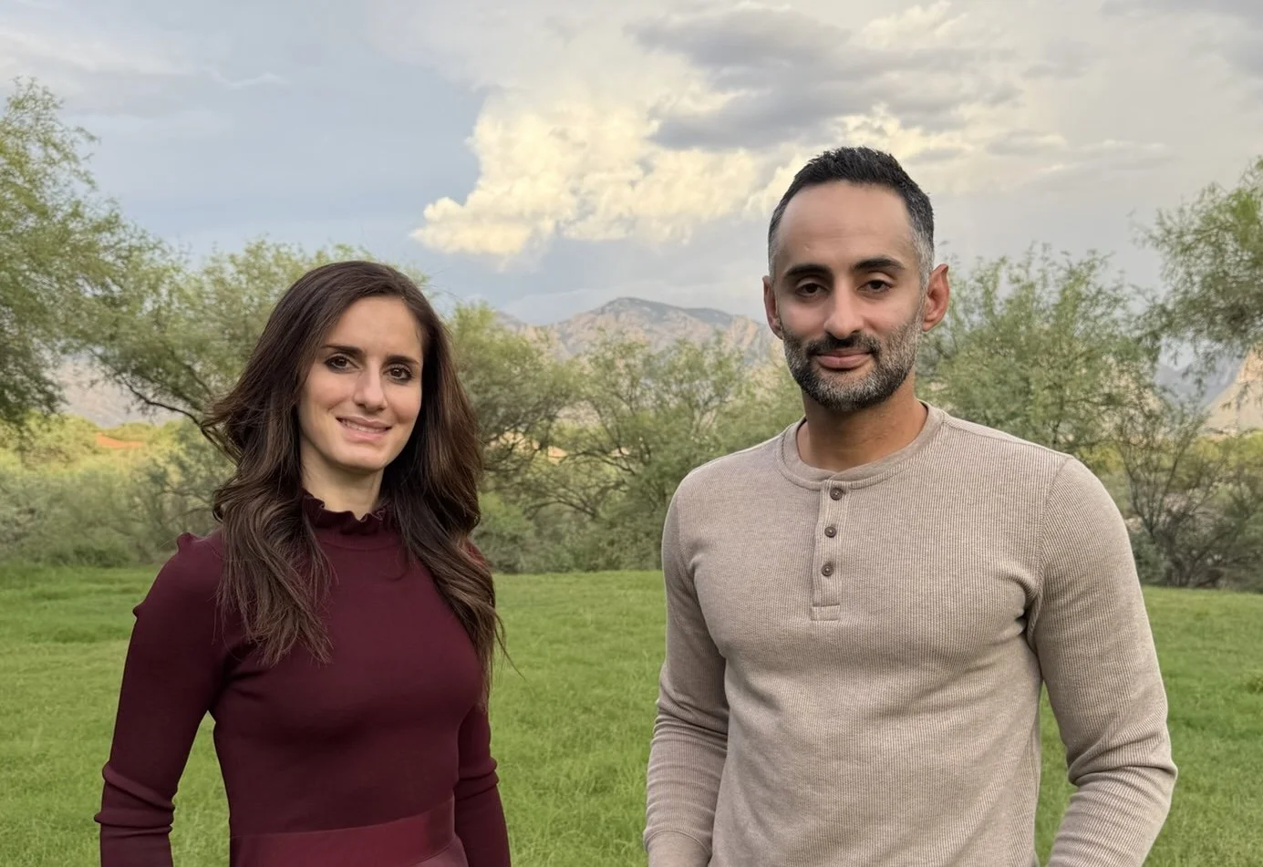 A woman with brown hair wearing a maroon long-sleeve top and a man with short dark hair and gray stubble wearing a beige long-sleeve shirt standing outdoors in a green field with trees and mountains in the background under a partly cloudy sky.