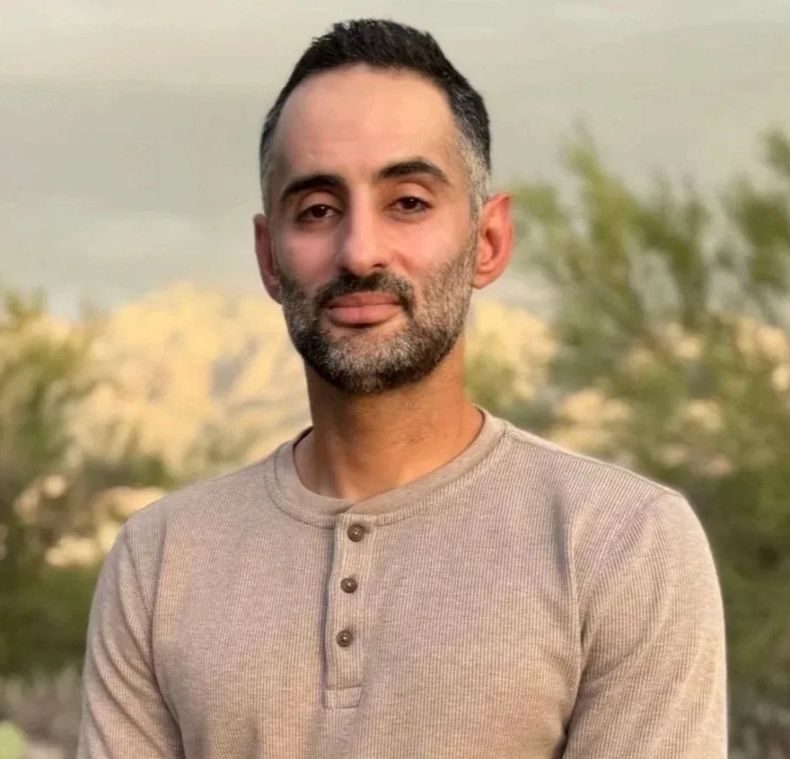 A man with short dark hair and a beard, wearing a beige Henley shirt, standing outdoors with trees and a cloudy sky in the background.