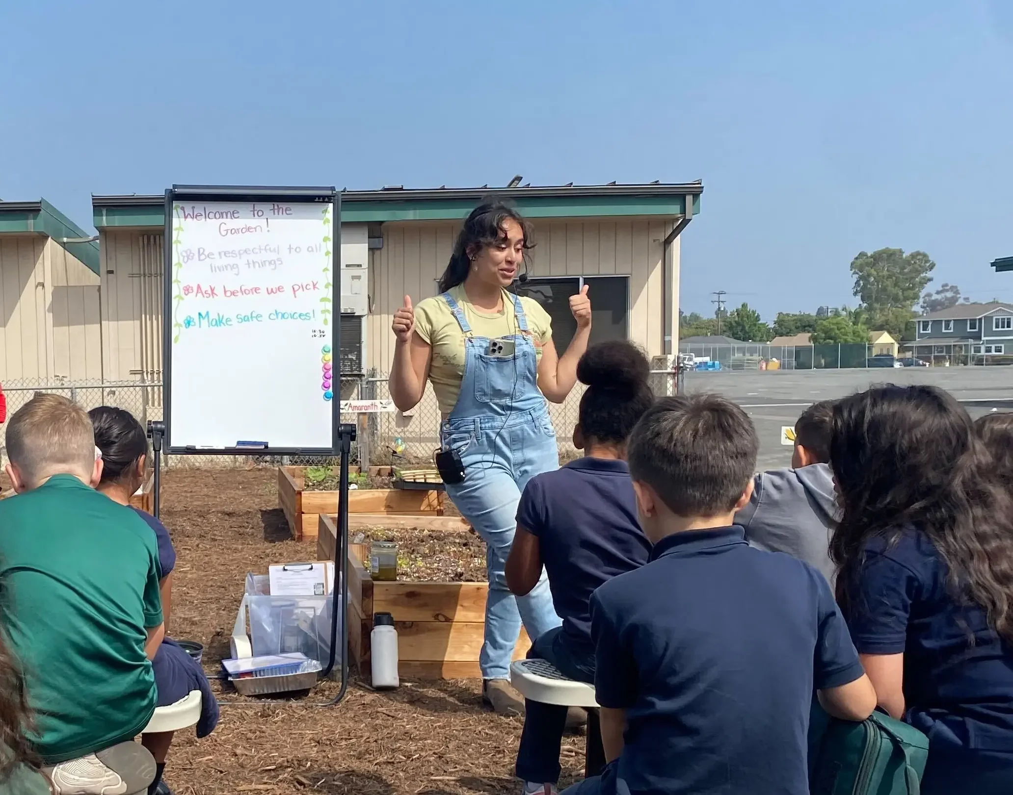 A Garden Educator teaches a class outside in the garden with a whiteboard behind her.