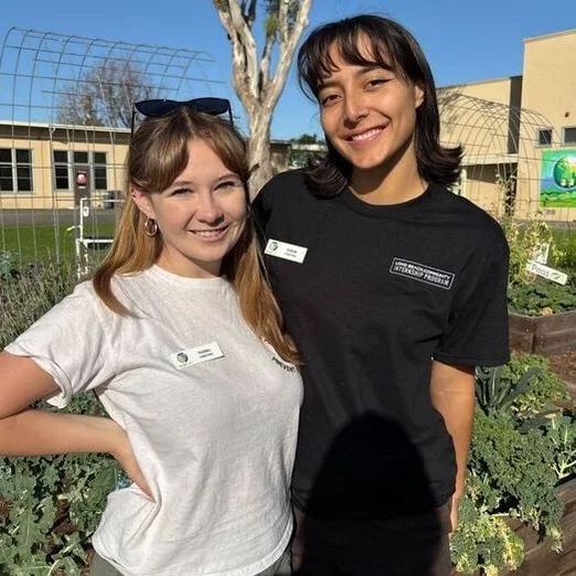 Two interns stand smiling in front of garden beds,