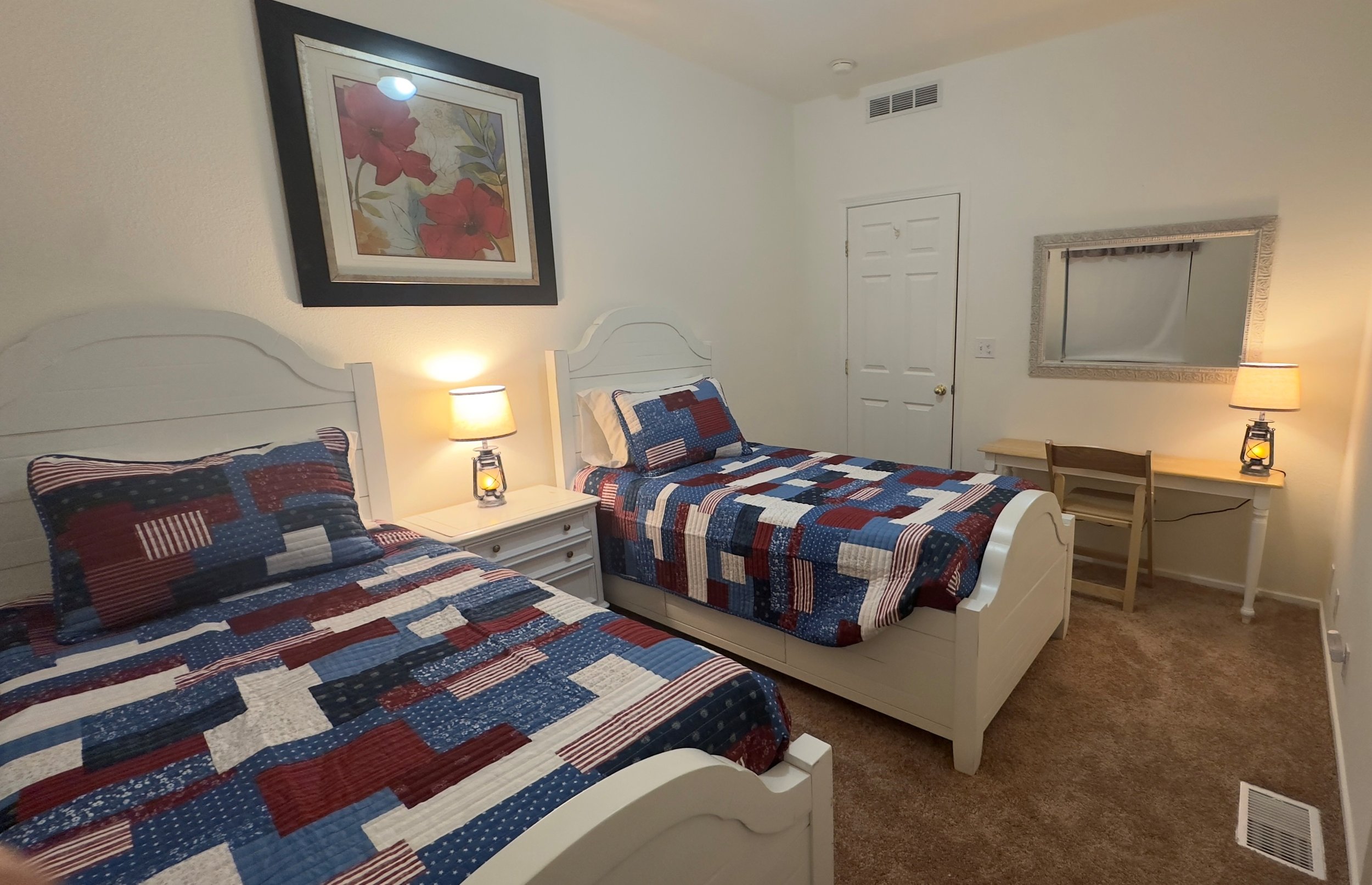 Two twin beds with red, white, and blue patchwork quilts in a bedroom. Each bed has a pillow with a matching quilt design. There are two nightstands with lamps, a wall painting of pink flowers, a mirror with a decorative frame, and a small desk with 