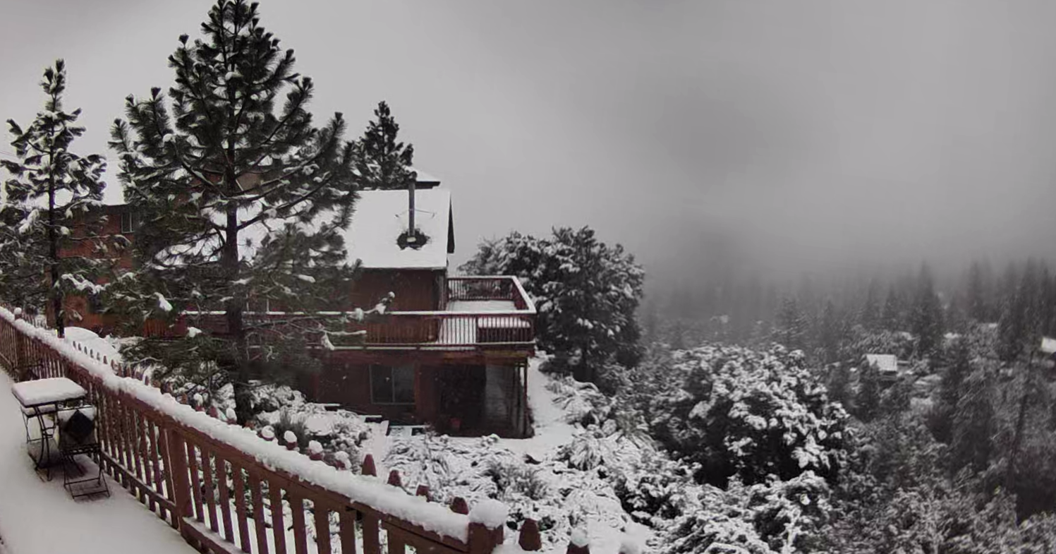 A snowy landscape with a wooden house and trees covered in snow, viewed from a balcony with snow-covered railing and furniture.