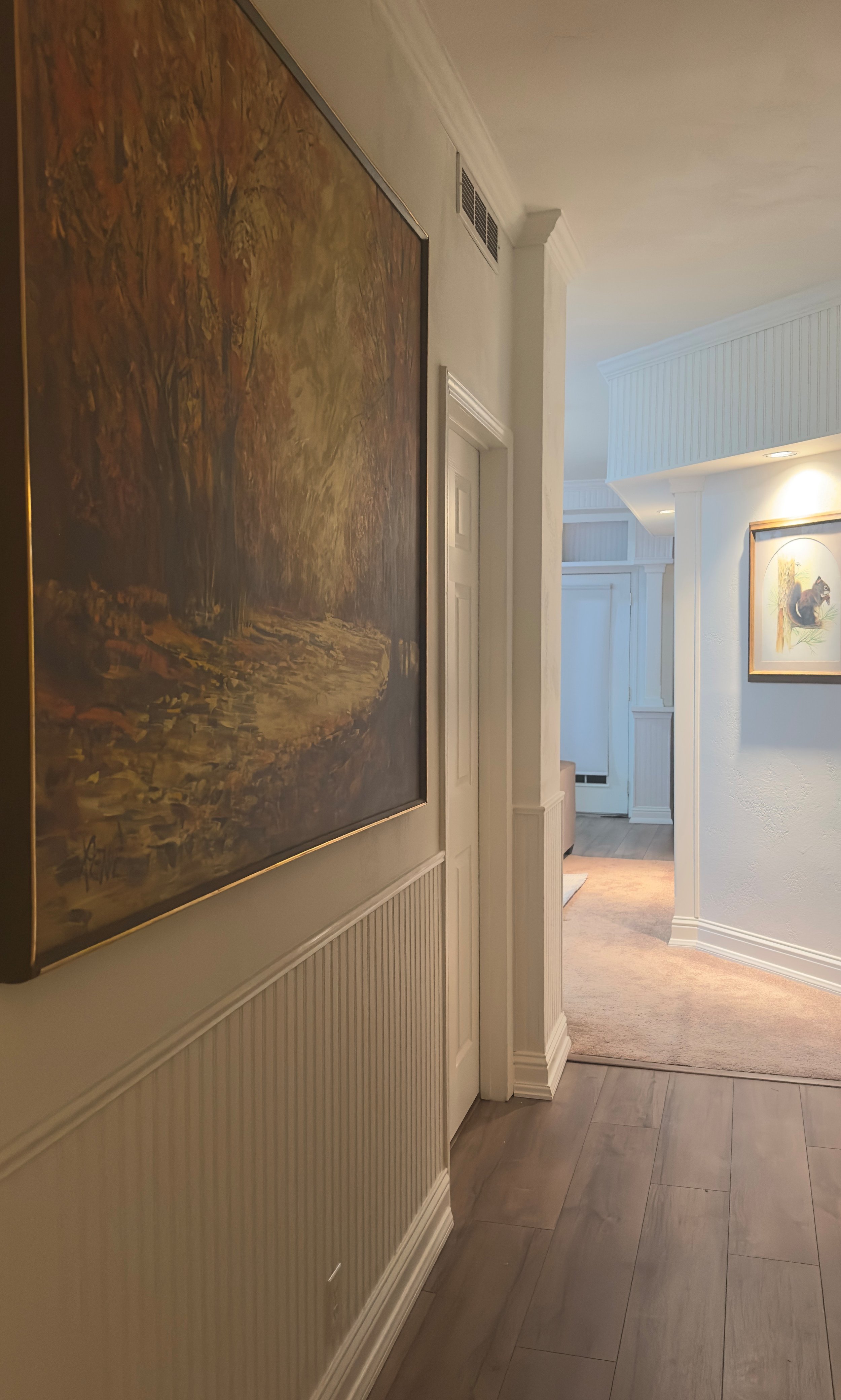 Image of an interior hallway with framed artwork on the walls, hardwood flooring, and an arch leading to another room with a carpet.