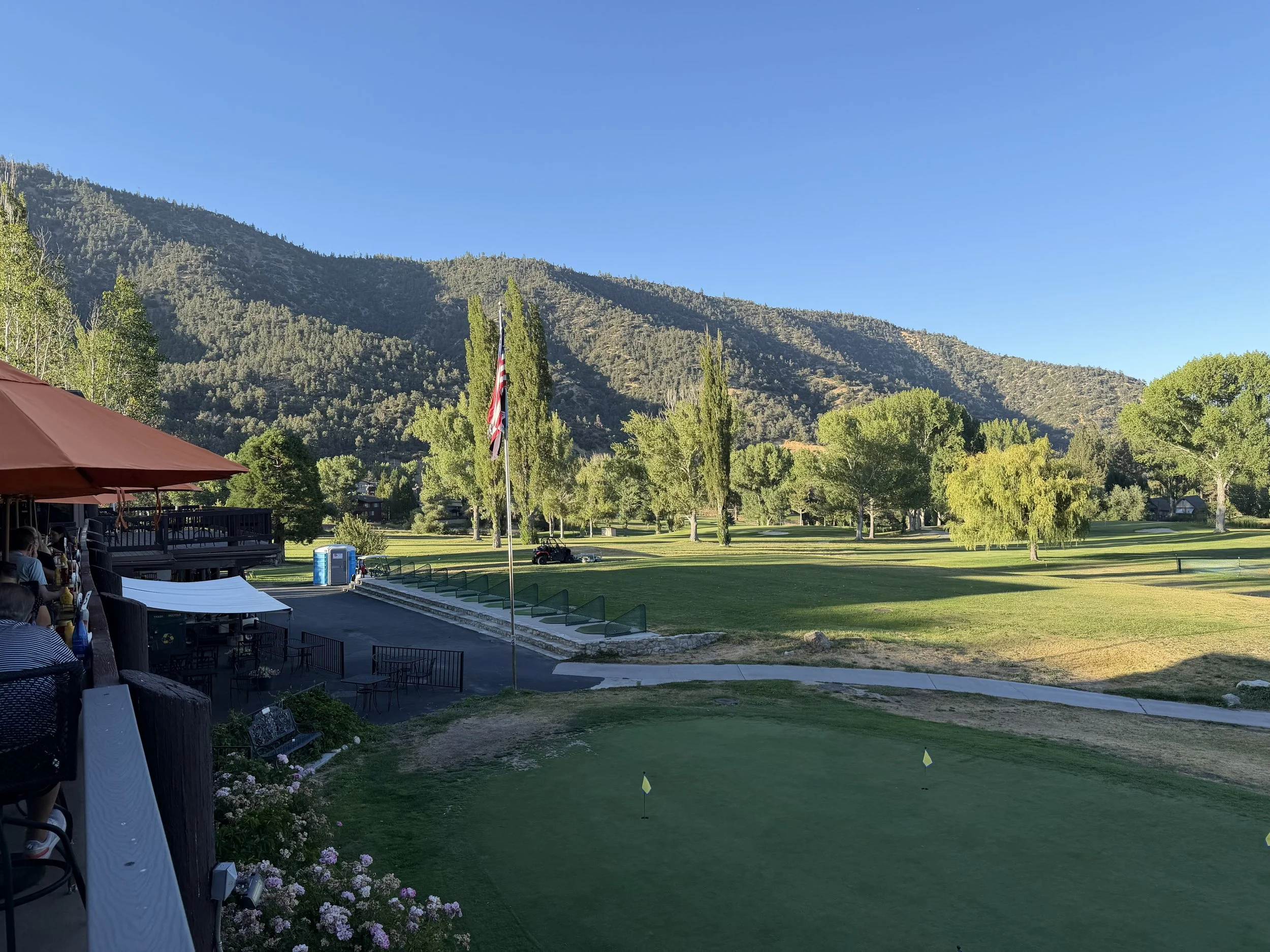 A scenic view of Pine Mountain Club Golf Course with a putting green in the foreground, flag markers, and surrounding trees. A flagpole with an American flag and caddy carts are visible. In the background, there are mountains under a clear blue sky.