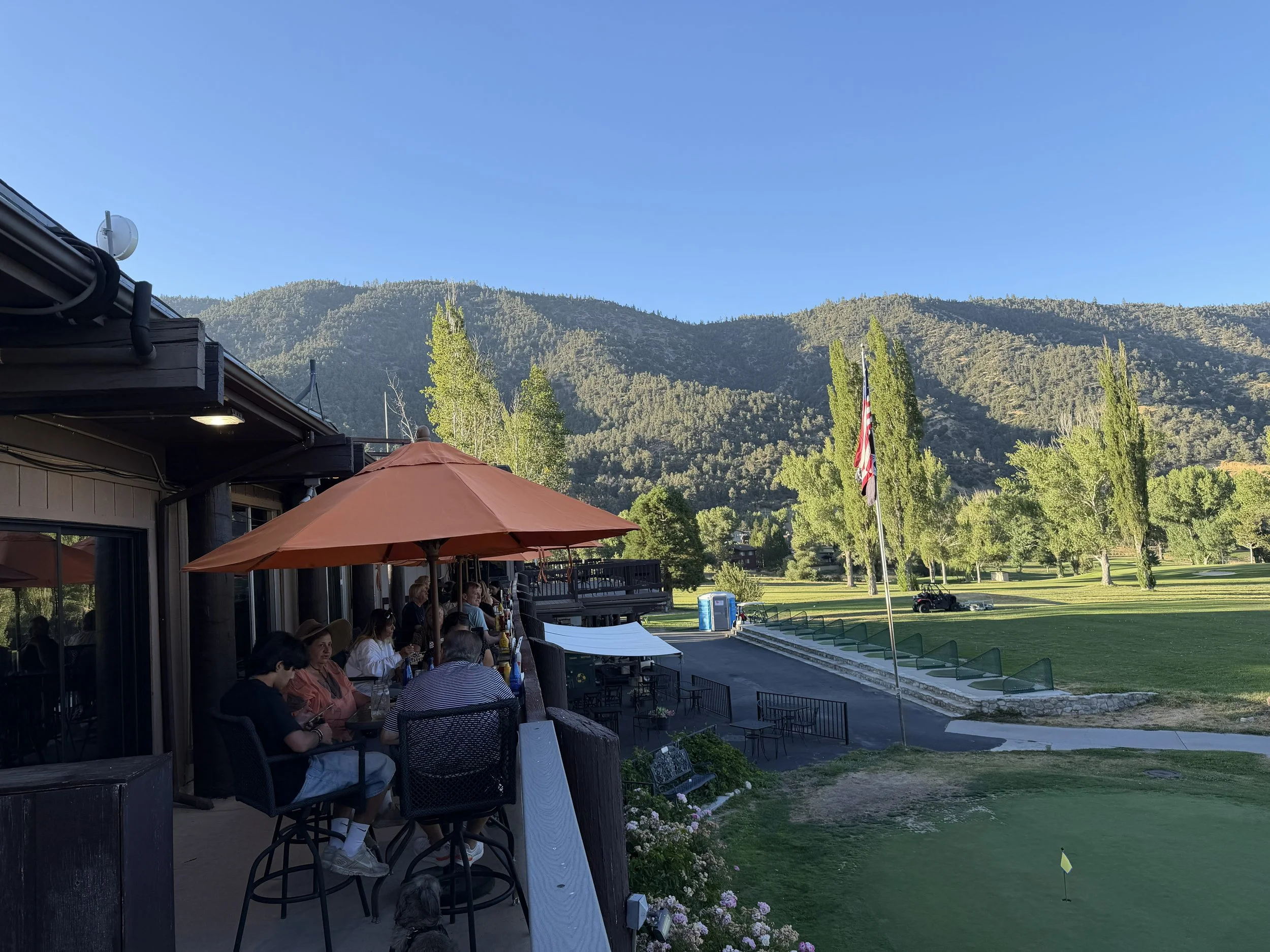 People are sitting at tables with orange umbrellas outside the Pine Mountain Club Golf Course with a view of green hills and a clear blue sky.