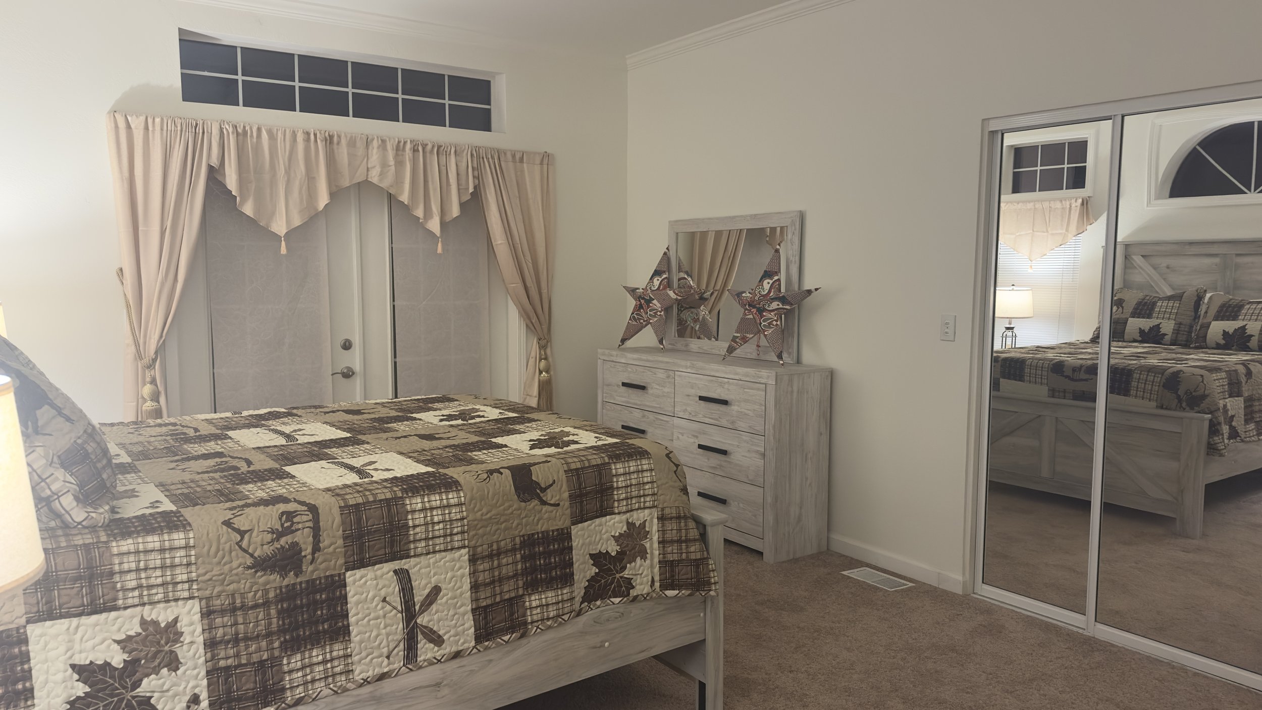 A bedroom with a bed featuring a plaid and leaf quilt, a white dresser with star decorations on top, a large mirror, and a mirrored closet door reflecting the room. There are beige curtains and a window above the door.