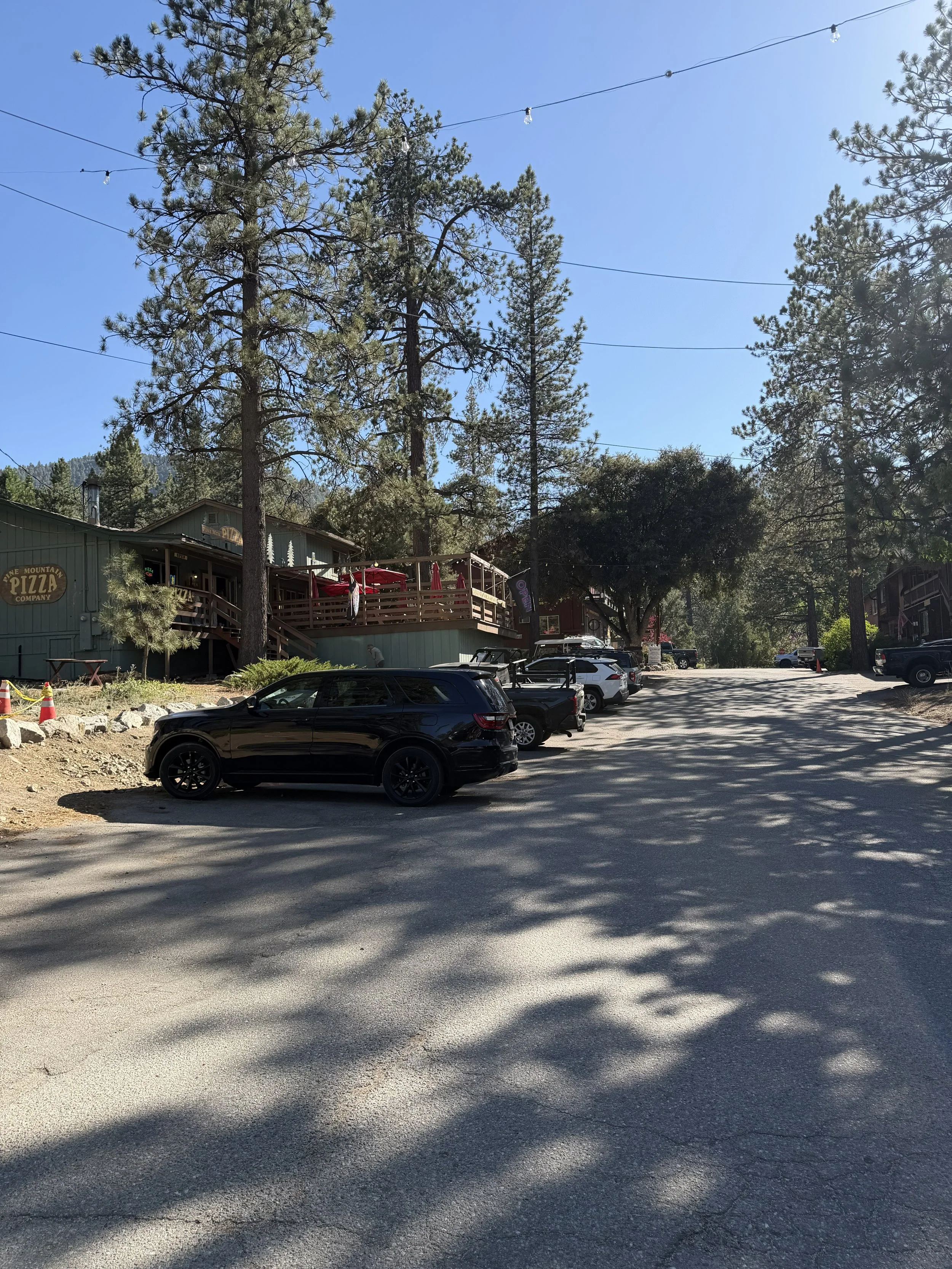A parking lot with several parked cars and a building in the background. Tall trees are visible, with power lines crossing the sky. The building has a sign that reads 'Pine Mountain Pizza Company' and a patio with red umbrellas.