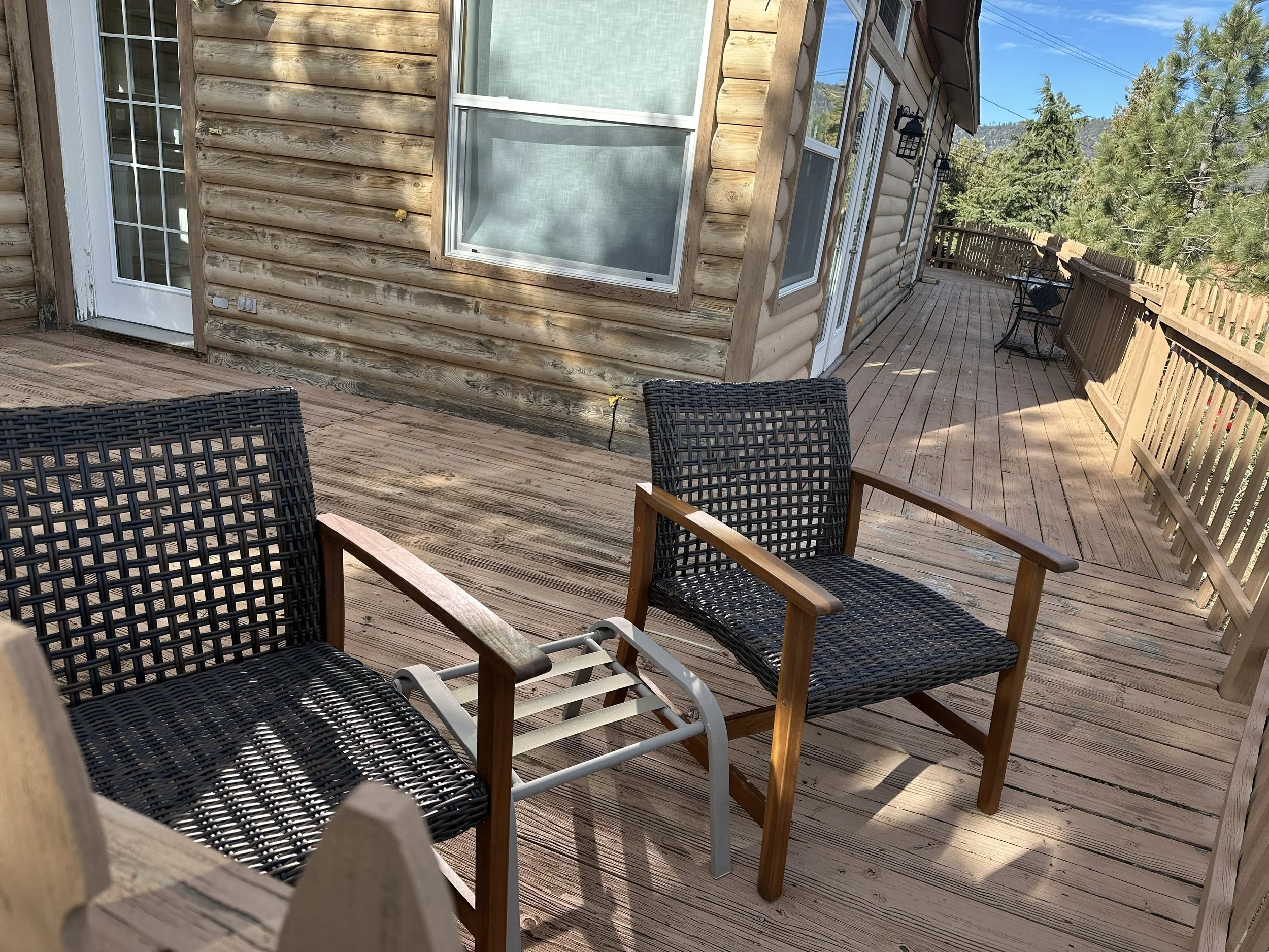 View of a wooden outdoor deck with black wicker chairs, a wooden bench, and a wooden fence. The house exterior has a wood siding and glass windows with a mountain and pine trees visible in the background.
