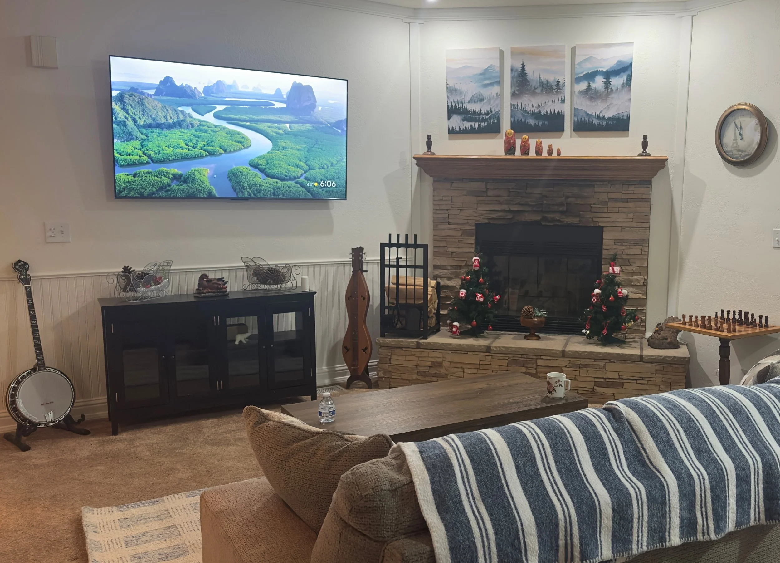 A living room with a wall-mounted flat-screen TV showing a scenic nature landscape with a river and lush greenery. To the right of the TV, there is a decorated fireplace mantle with artwork depicting mountains and forests, small decorative figurines,