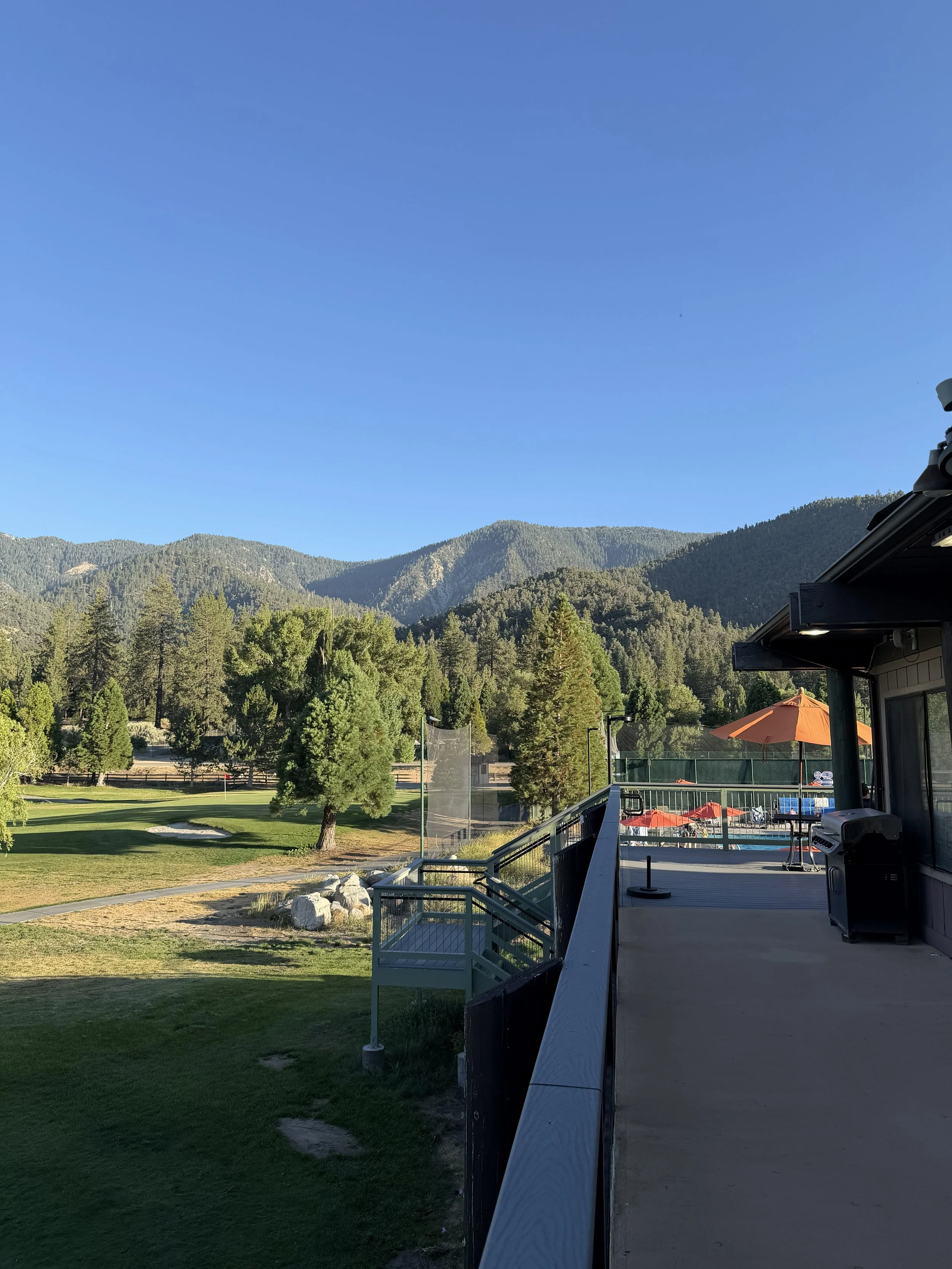 The Pine Mountain Clubhouse with a view of green mountains under a blue sky. There is a patio with an orange umbrella, a grill, and a railing, along with some trees and a golf course.