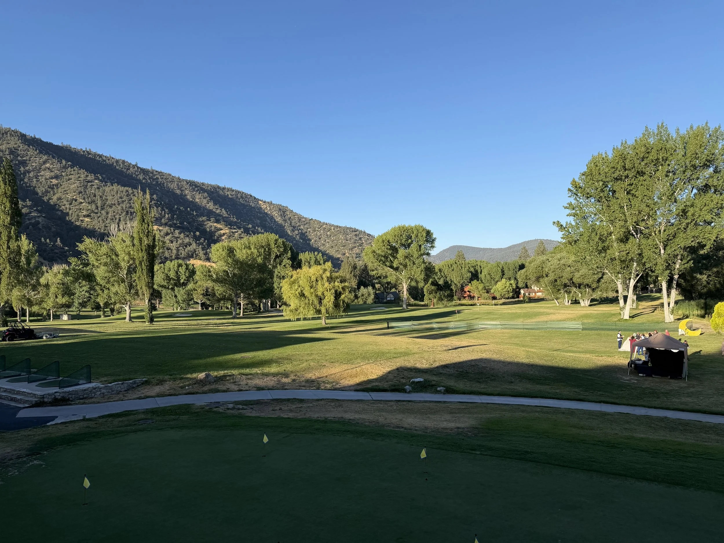 Pine Mountain Club Golf Course with green grass, trees, mountains in the background, and a clear blue sky. There are small flags marking holes and a few people near a tent on the right side.