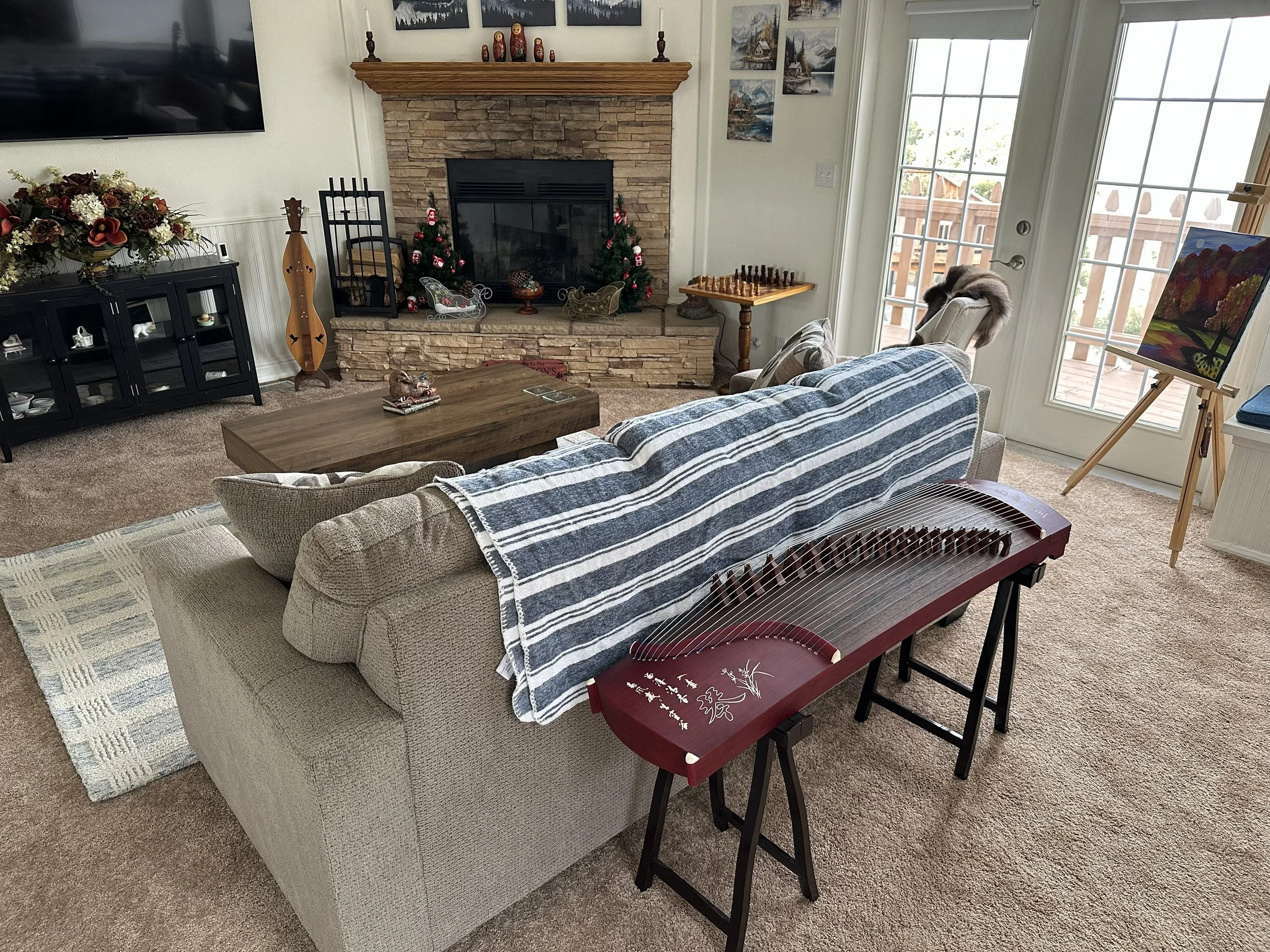 Living room with a beige couch, a wooden coffee table, and a striped blanket. A fireplace with Christmas decorations and a small tree is in the background. A black cabinet with floral arrangements is on the left, and an art easel with a painting is n