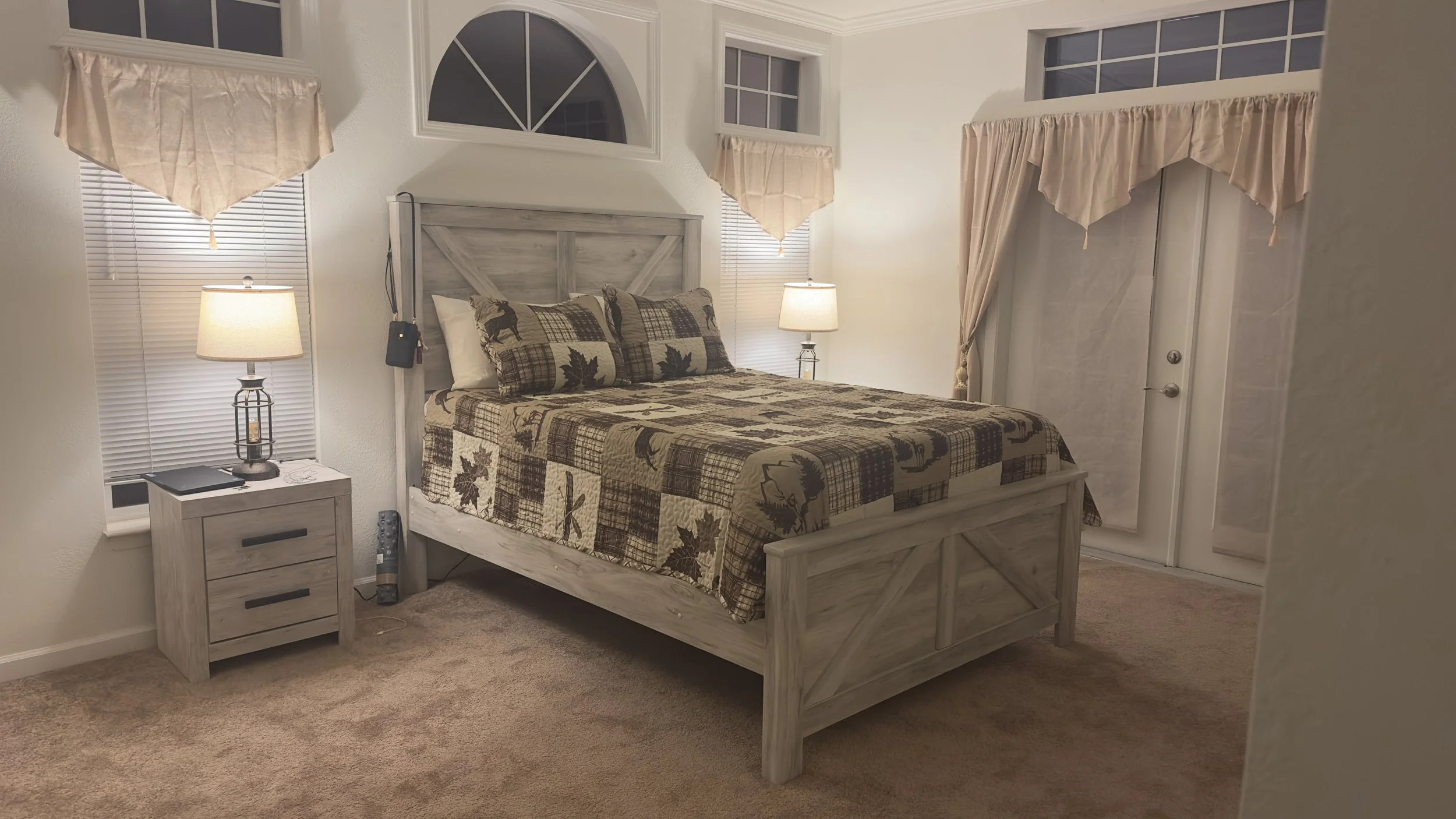 A bedroom with a wooden bed frame, plaid and leaf-patterned bedding, and pillows. There are two nightstands with lamps on each side of the bed. The room has white walls, beige carpet, and small windows with beige curtains.