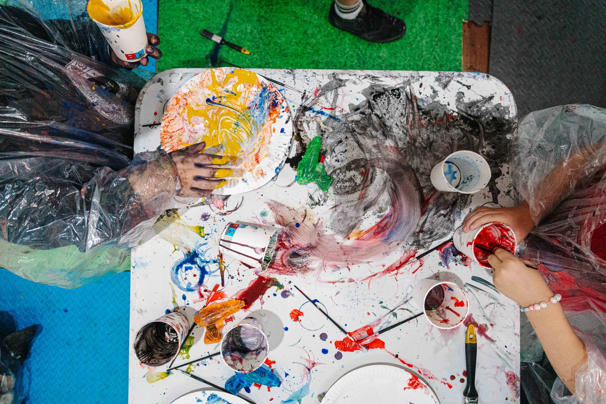 Children and adults creating abstract paintings with bright colors on paper and a messy table with paint containers, brushes, and other art supplies.