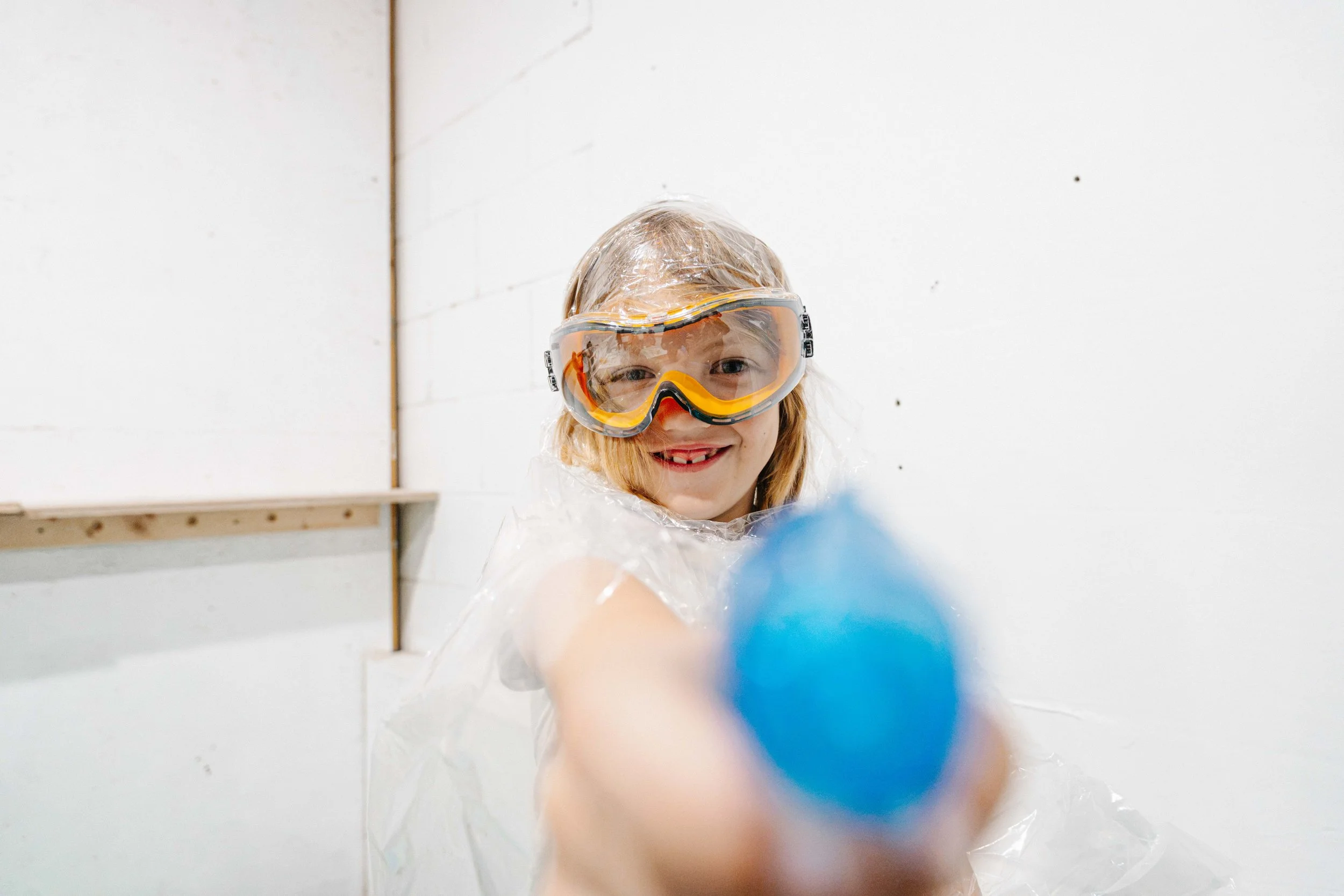 A child wearing safety goggles smiles while reaching towards the camera, with a white wall background.