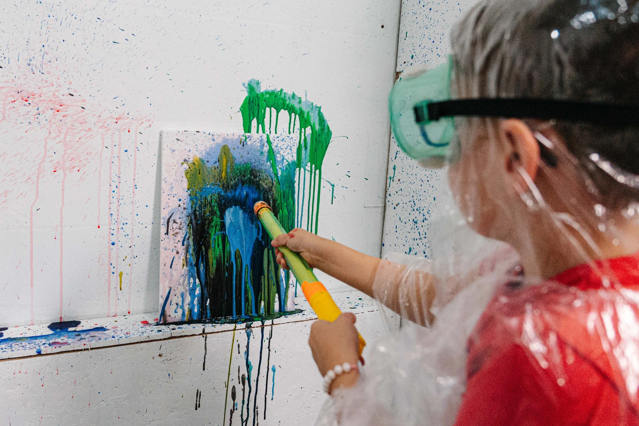 A child wearing safety goggles and a red paint-splattered smock is creating abstract art on a white canvas using vibrant colors including green, blue, and yellow, with paint drips and splatters around.