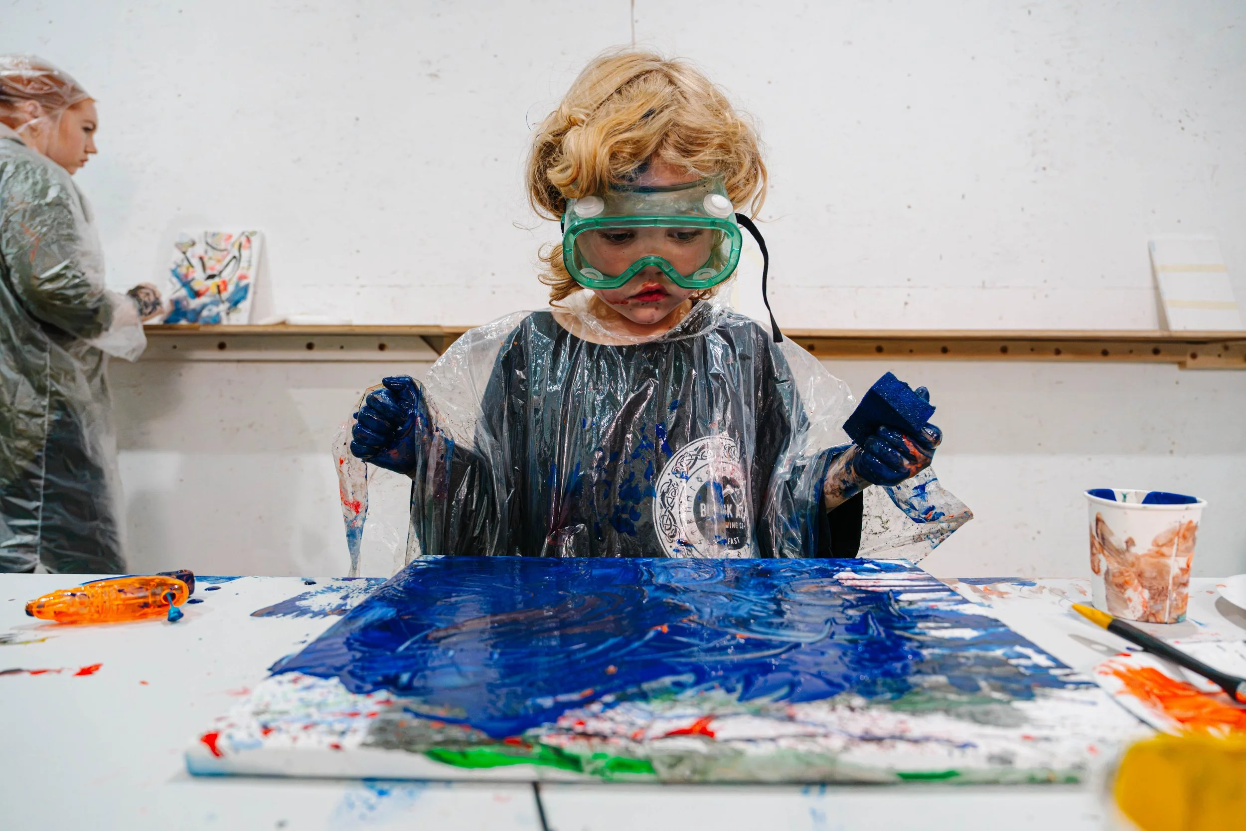 A young child with curly blonde hair wearing a green goggles, a plastic smock, and blue gloves, engaged in finger painting with blue paint on a table in an art classroom.