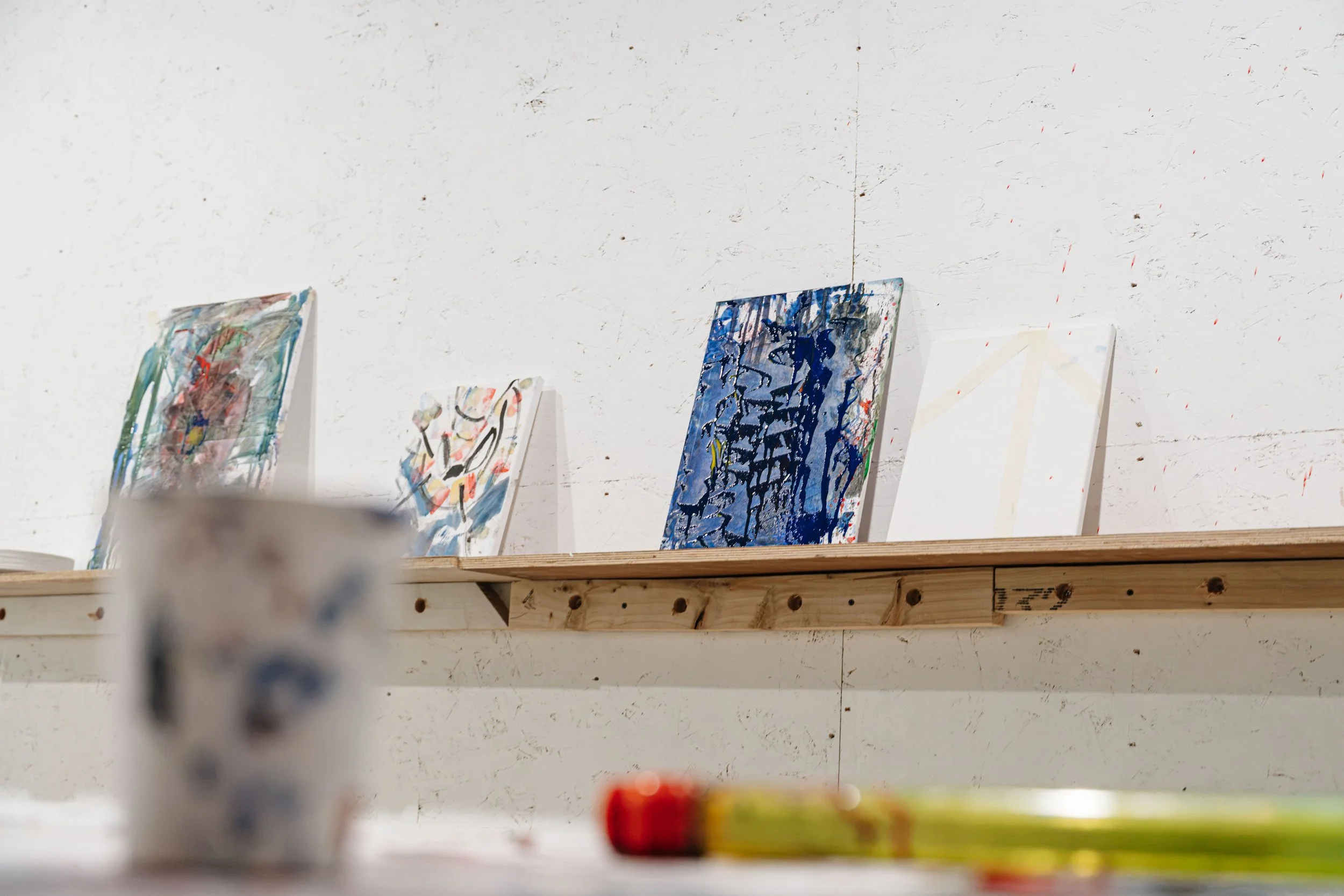 Several abstract paintings on canvases displayed on a wooden shelf against a white wall with red paint splatters.