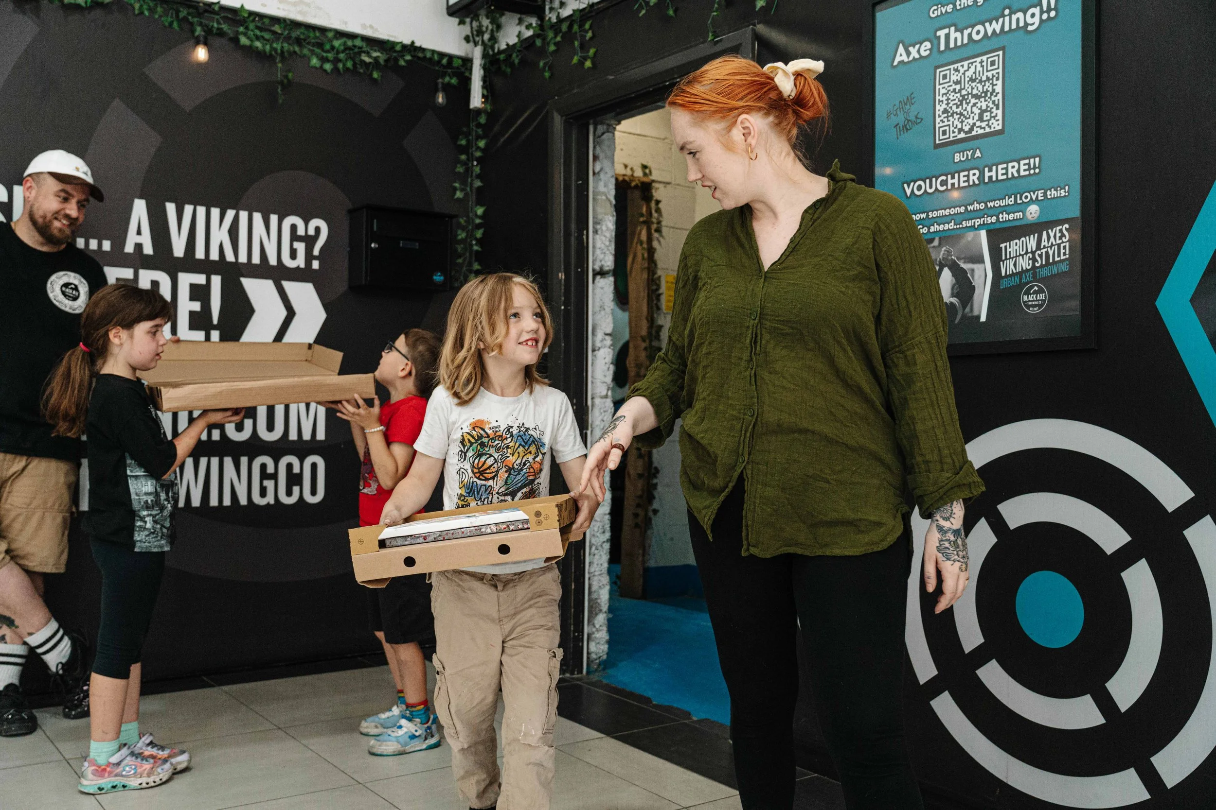 A woman with red hair and tattoos on her arm is standing in front of children receiving pizza boxes from a young girl at an indoor event. The girls are lined up, and there is a man smiling in the background. The scene appears to be at a fun activity or arcade, with signs advertising axe throwing.