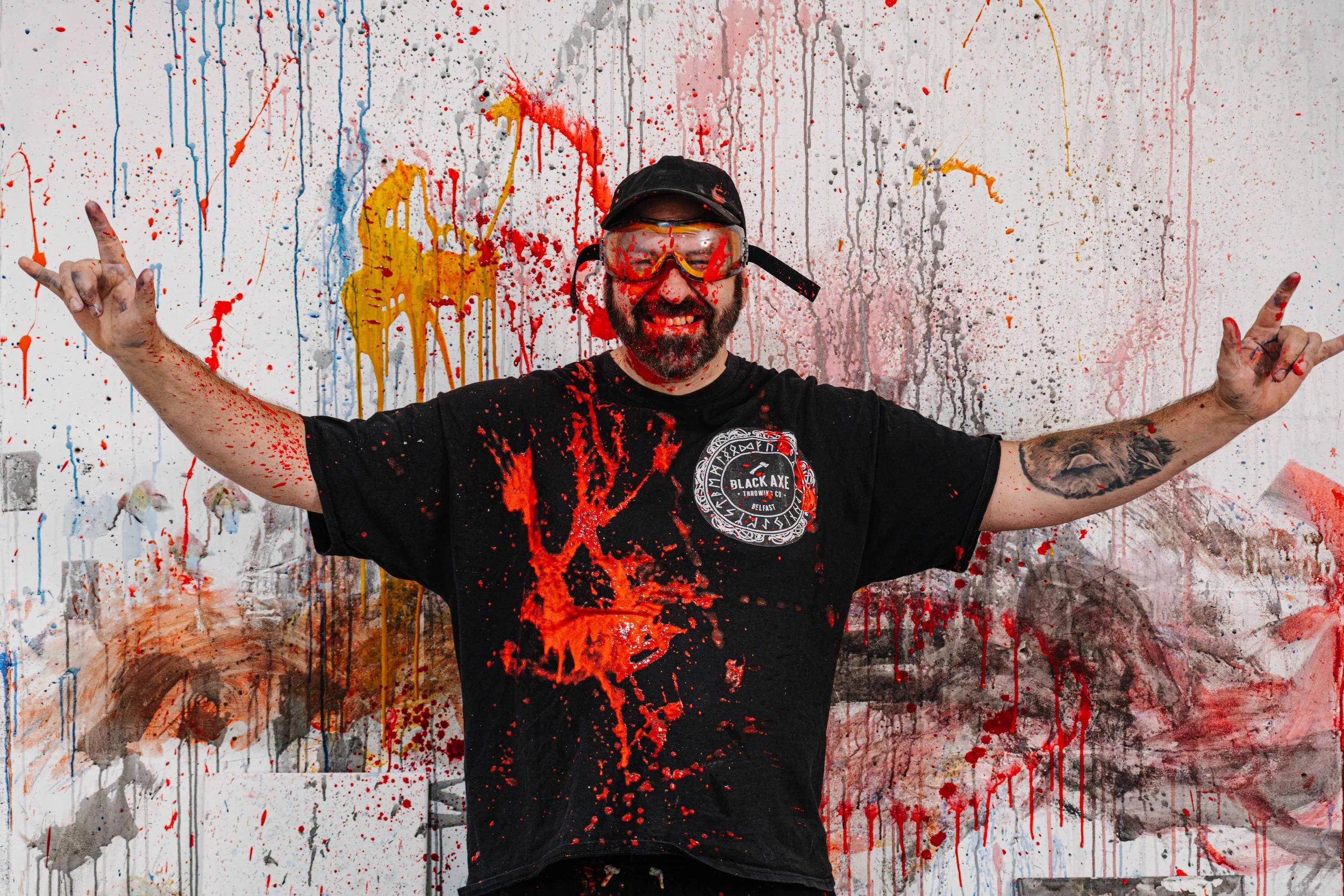 A man with a beard in black clothing and goggles stands in front of a wall covered in colorful paint splatters and drips, smiling with arms outstretched.