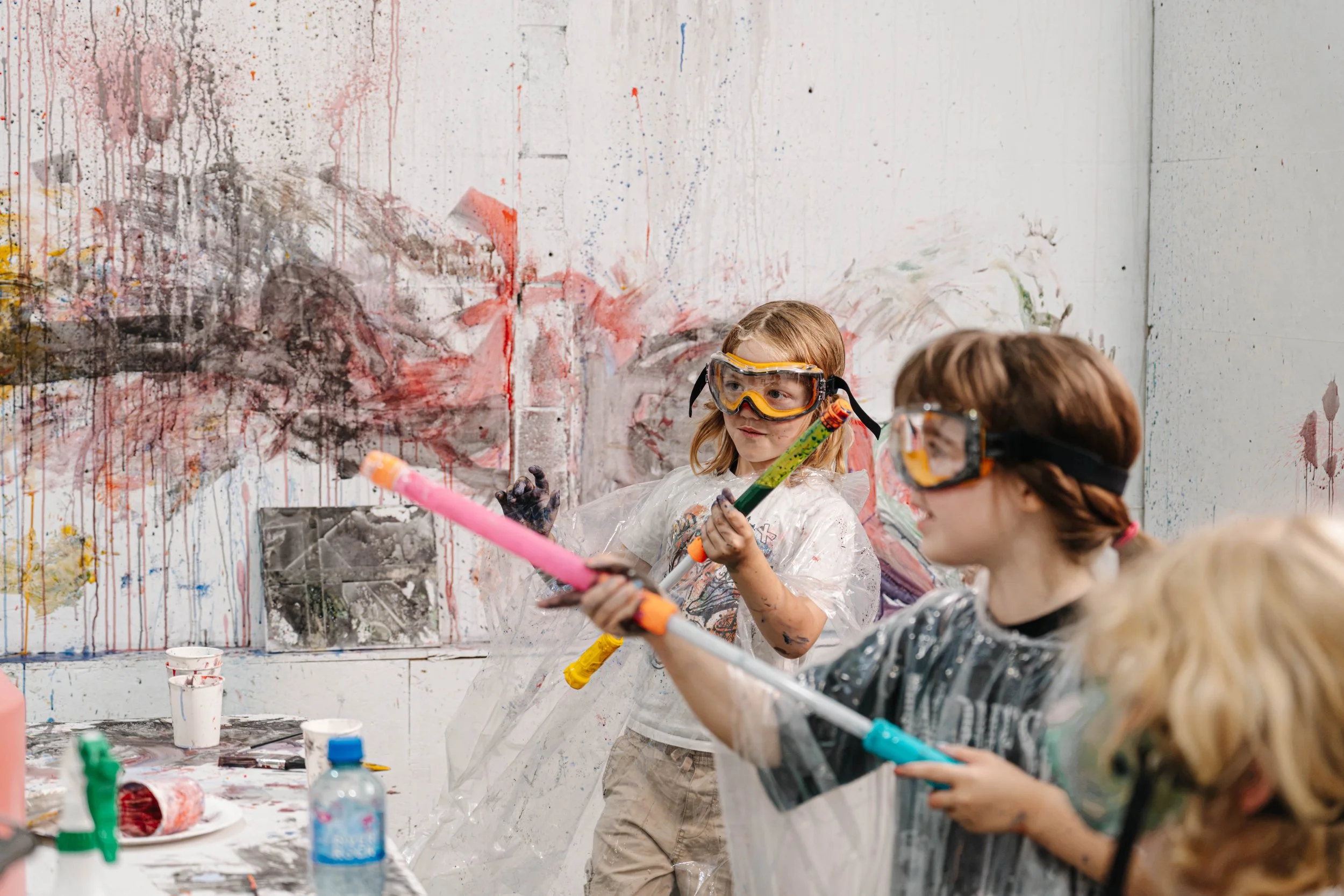 Kids wearing safety goggles painting and creating art on a messy white wall with splashes of paint, paintbrushes, and cups of paint on a table.
