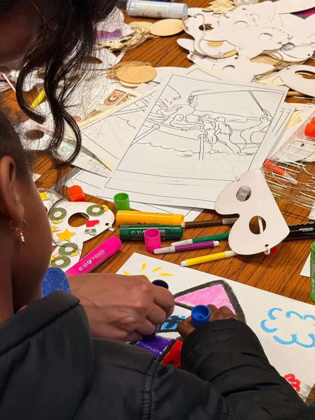 Children create Christmas-themed art with coloring supplies, including markers, stickers, and paper masks with holiday decorations.