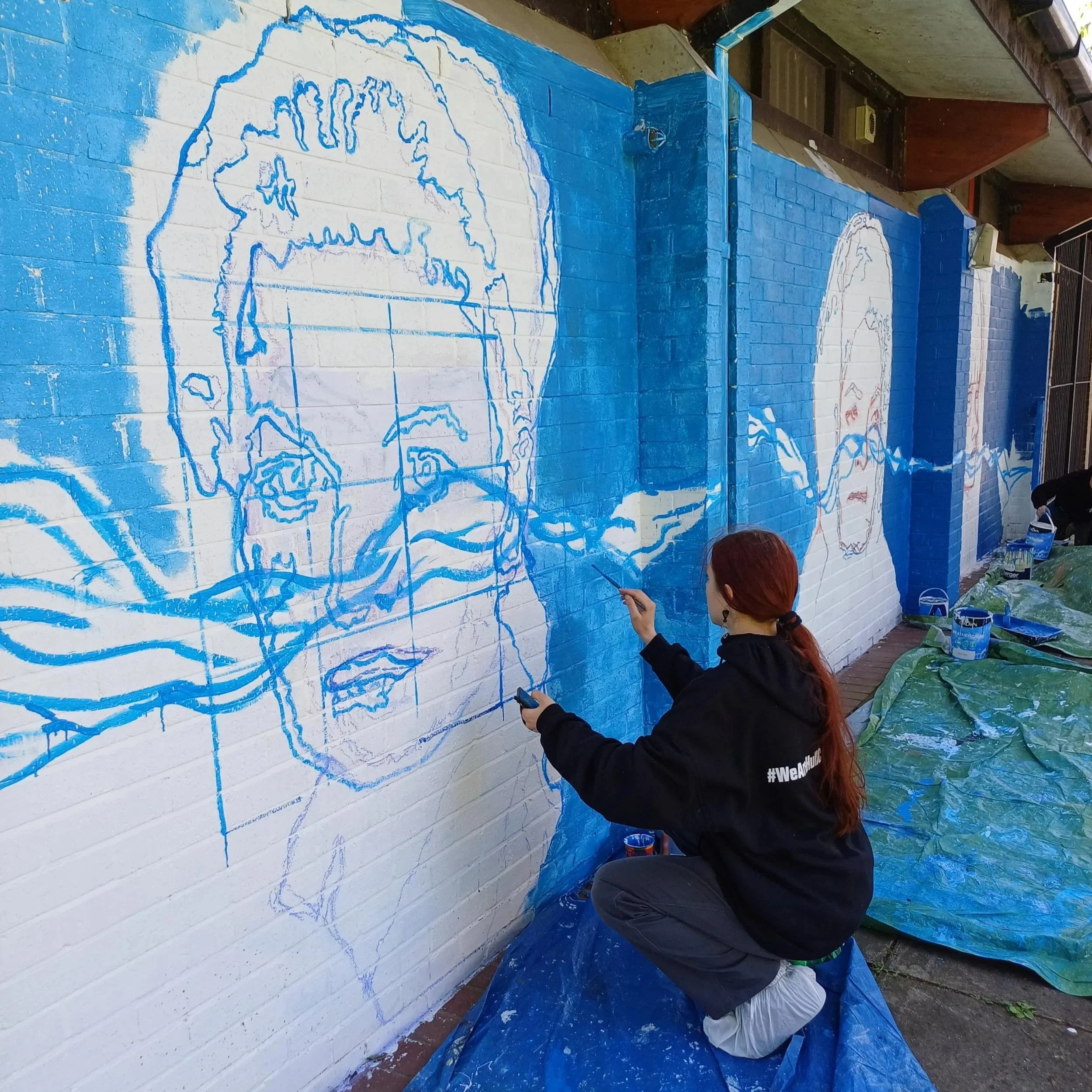 A person with red hair painting a mural on a brick wall with blue paint. The mural depicts a map outline of a region with some details inside. The person is wearing a black hoodie with white text on the sleeve and is seated on the ground while applyi