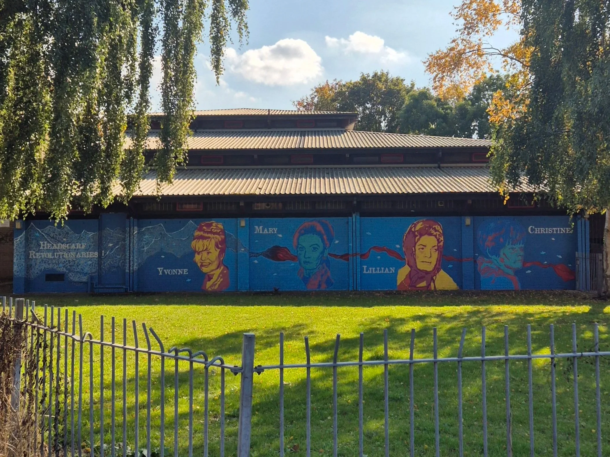 A colorful mural on a fence depicts various faces with names, set against a background with mountains and text, with a green grassy area and a small white fence in the foreground. The building behind the mural has a grey roof and trees with autumn fo
