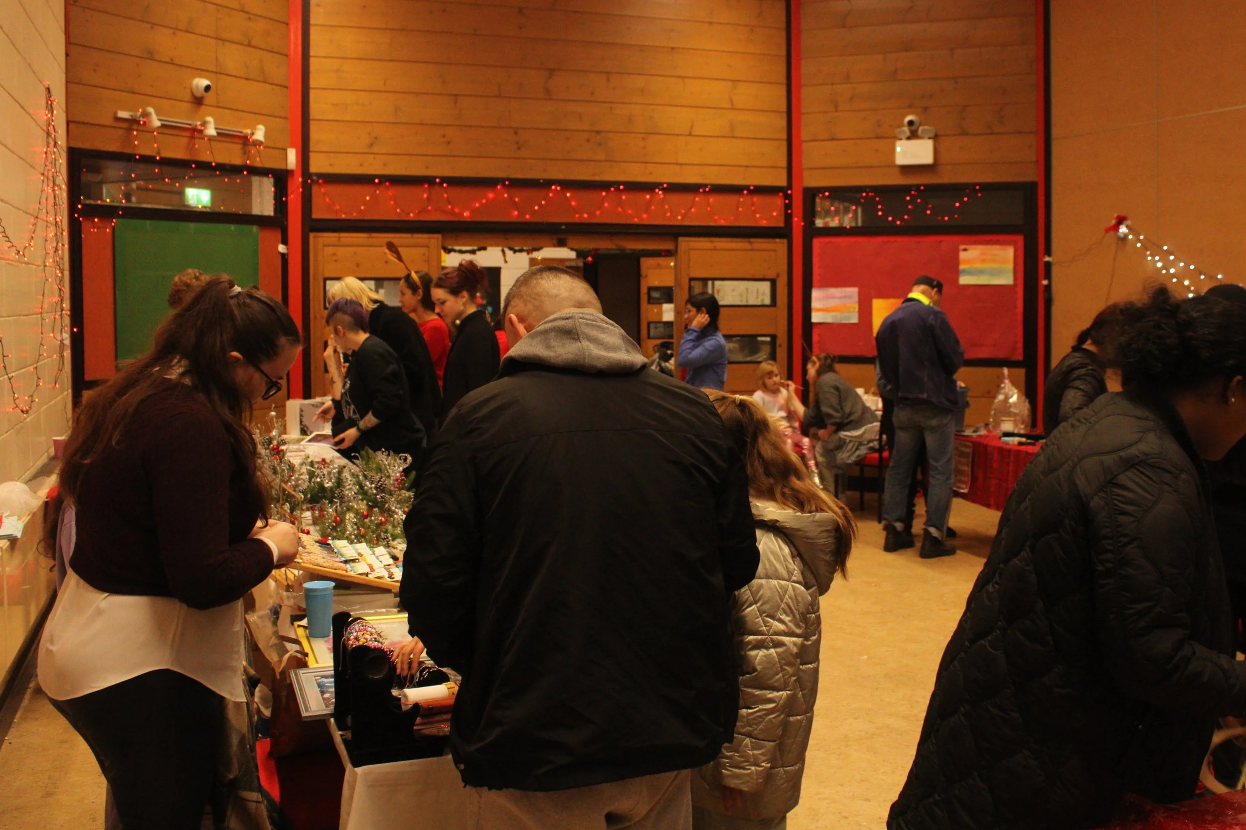 Indoor holiday market with people browsing tables of crafts and decorations, Christmas lights hanging on wooden walls.