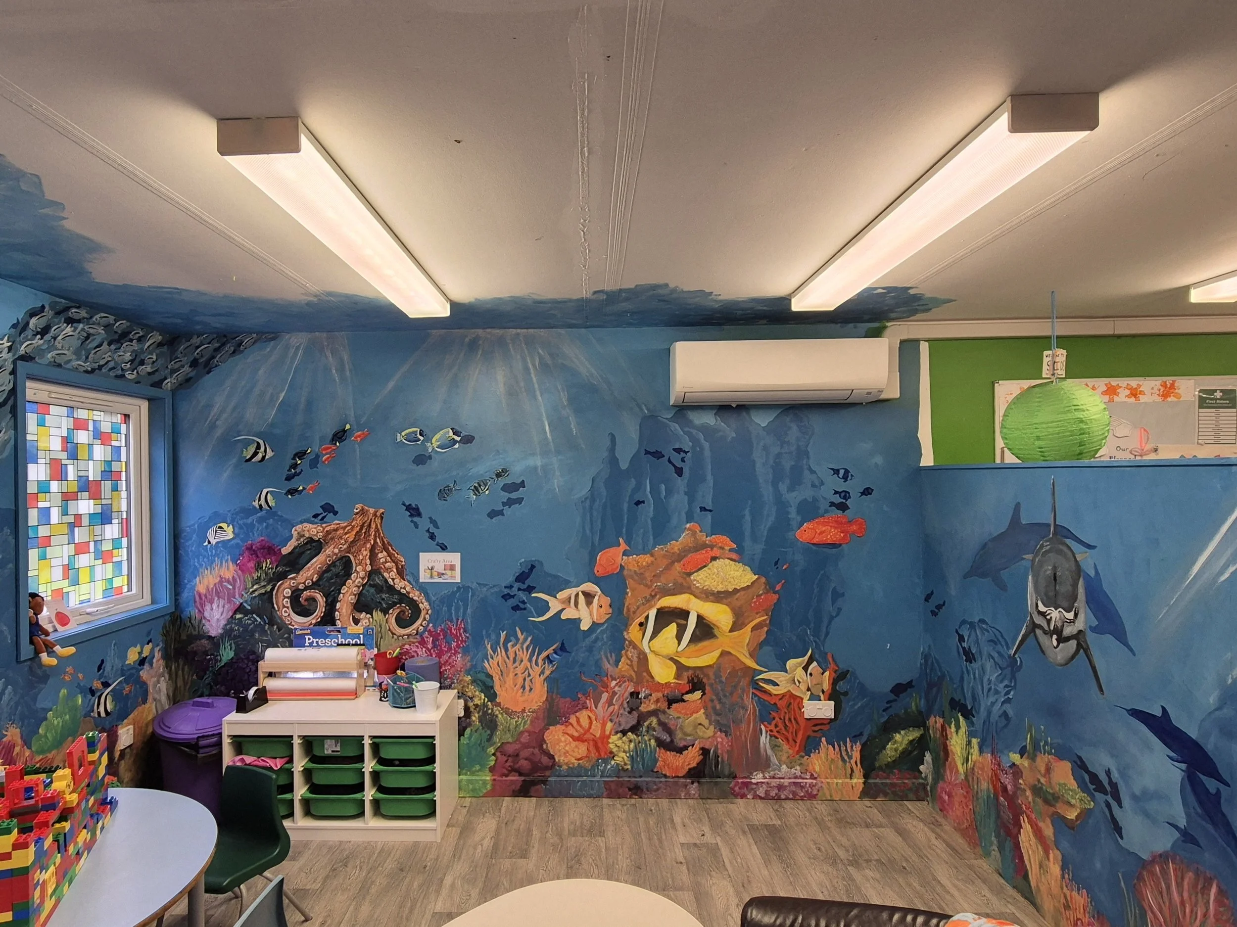 A colorful underwater mural on a classroom wall featuring various fish, coral, and sea creatures. The room has child-sized furniture, toys, and educational materials, indicating a preschool setting.