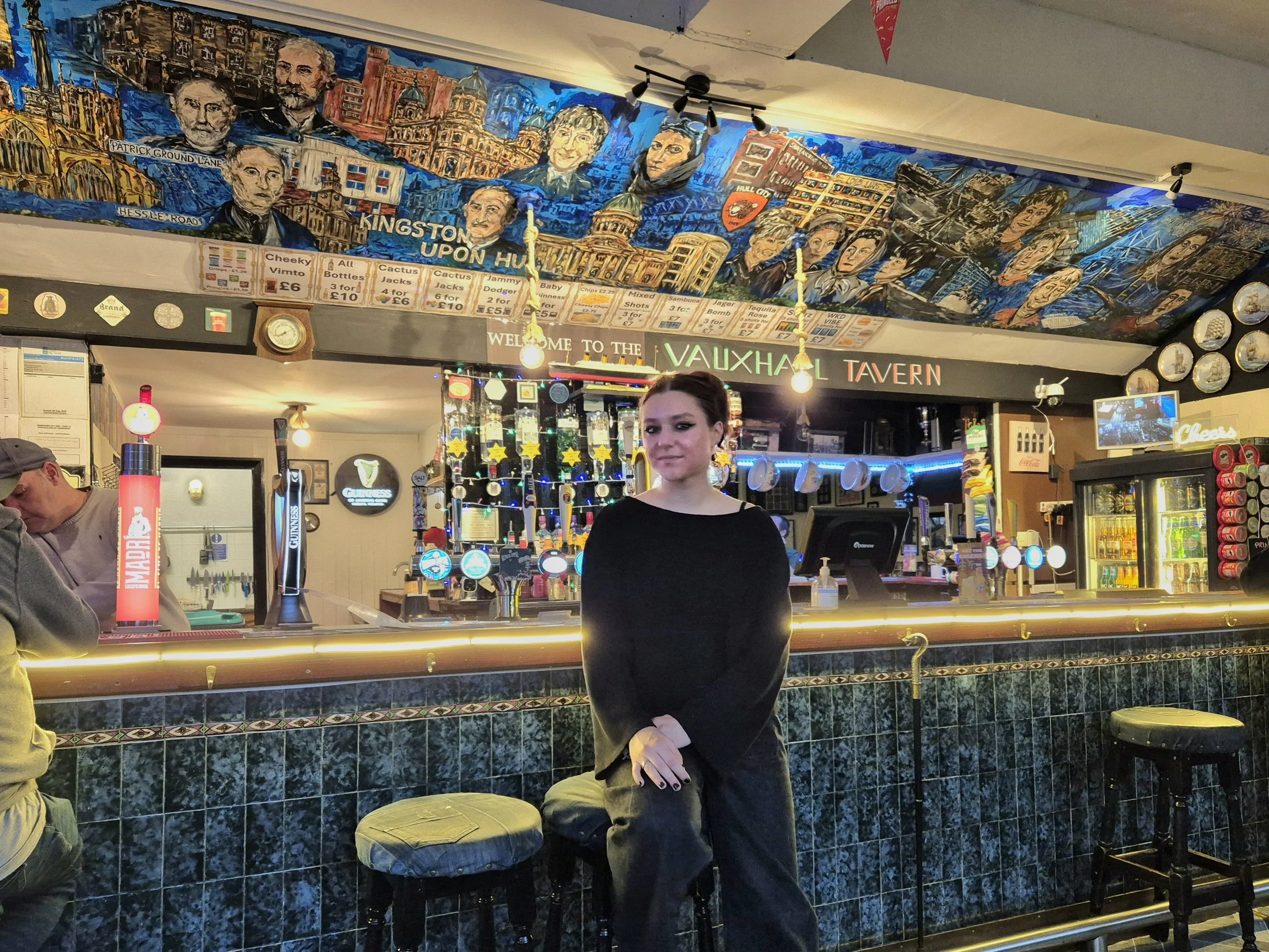 A woman stands behind a bar counter inside a pub or tavern, with colorful artwork and murals of buildings and portraits painted on the wall behind her. Various bar drink taps and bottles are visible, as well as hanging glasses and decorative lights.