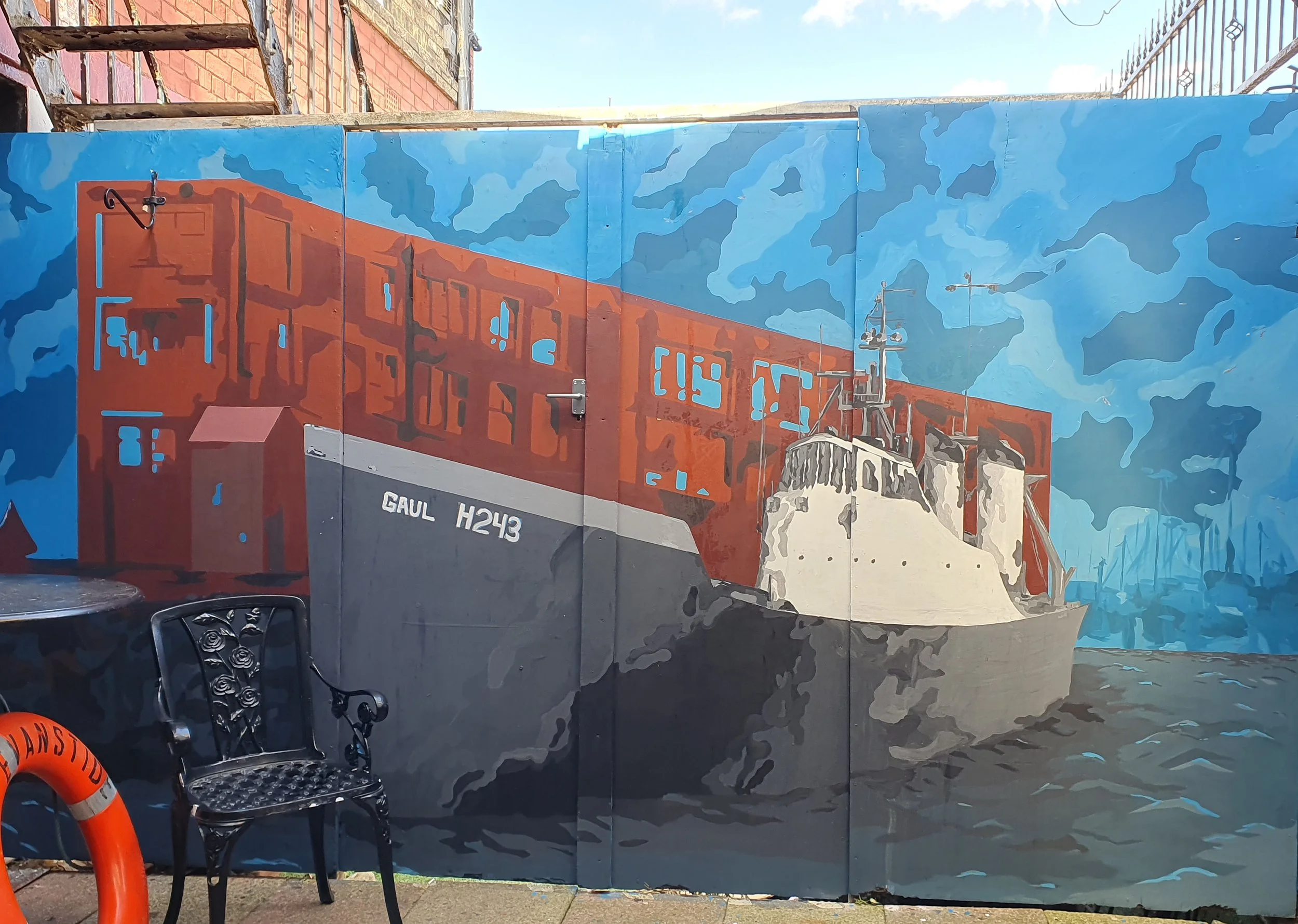 A large mural painted on a wall depicts a city scene with a building, a church, and a sky with scattered clouds. There is outdoor furniture including metal chairs and a lifebuoy at the bottom left of the image.