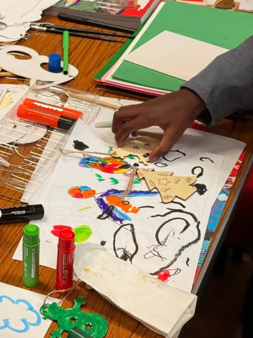 A table covered with art supplies and colorful drawings, with a person's hand reaching toward the paper with wooden craft pieces.