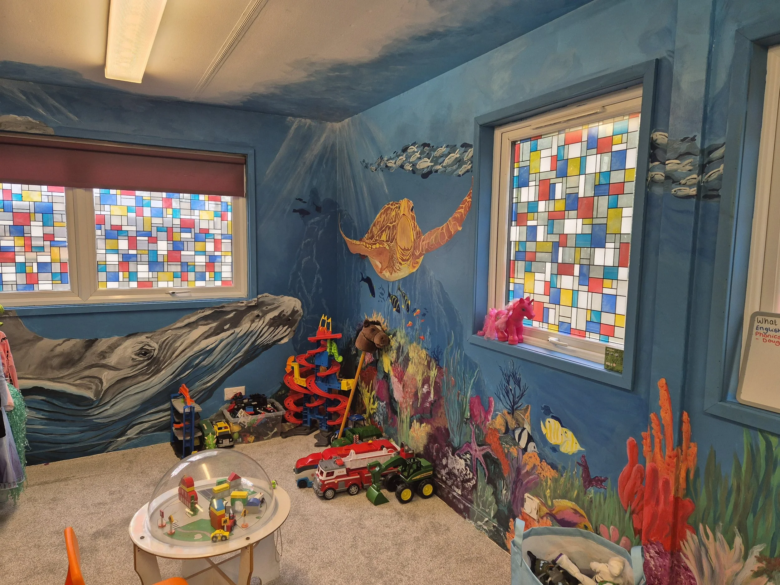 A colorful children's playroom with a mural of an underwater scene, including a large orange fish, smaller fish, and water plants. The room has stained glass windows depicting small, multi-colored rectangles, and various toys such as toy cars and a p