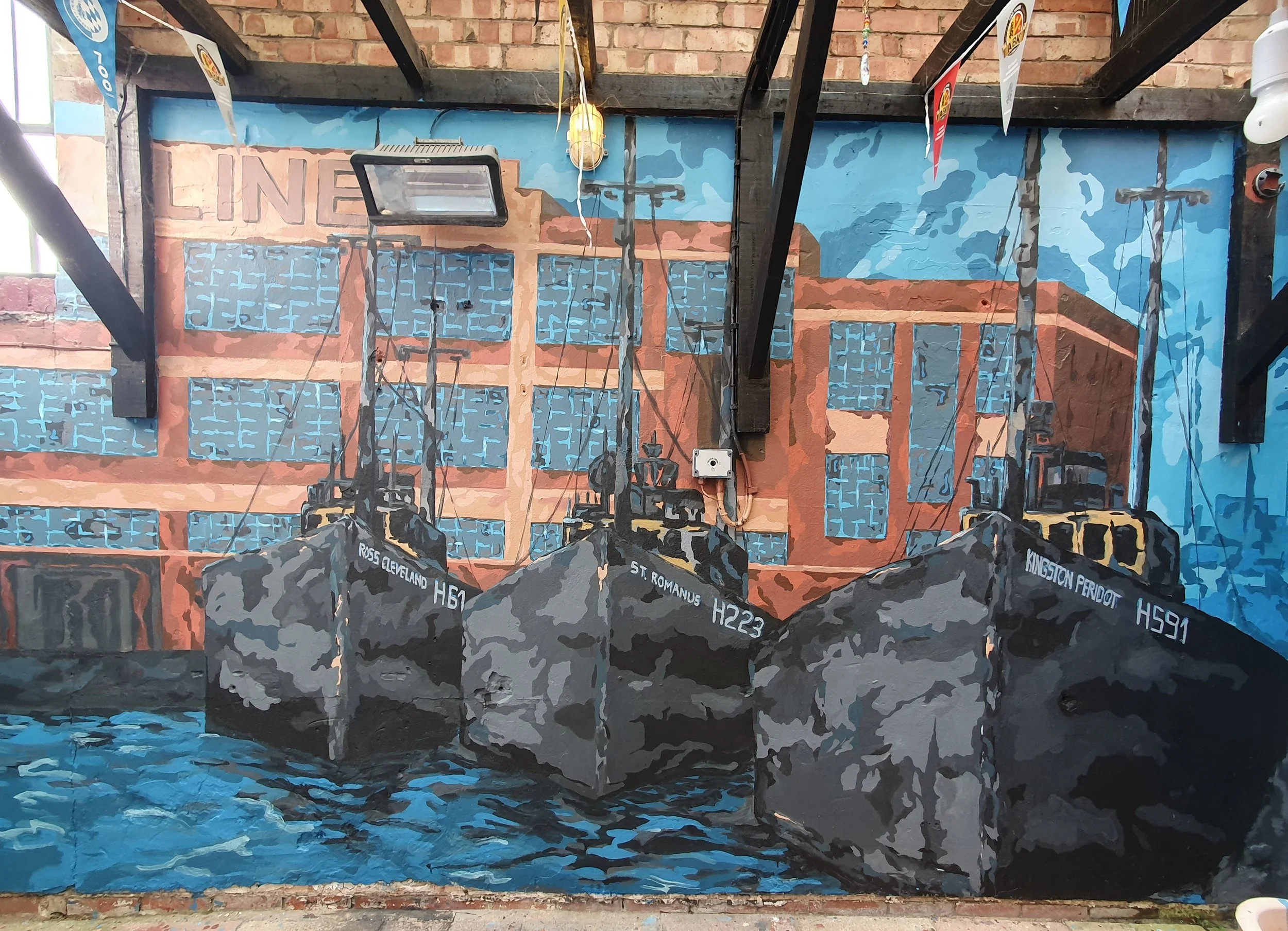 A mural painted on a brick wall features a cityscape with blue windows and orange building outlines, and a body of water in the foreground. Various pipes and wires are attached to the wall, with a light fixture hanging from above. There are several f