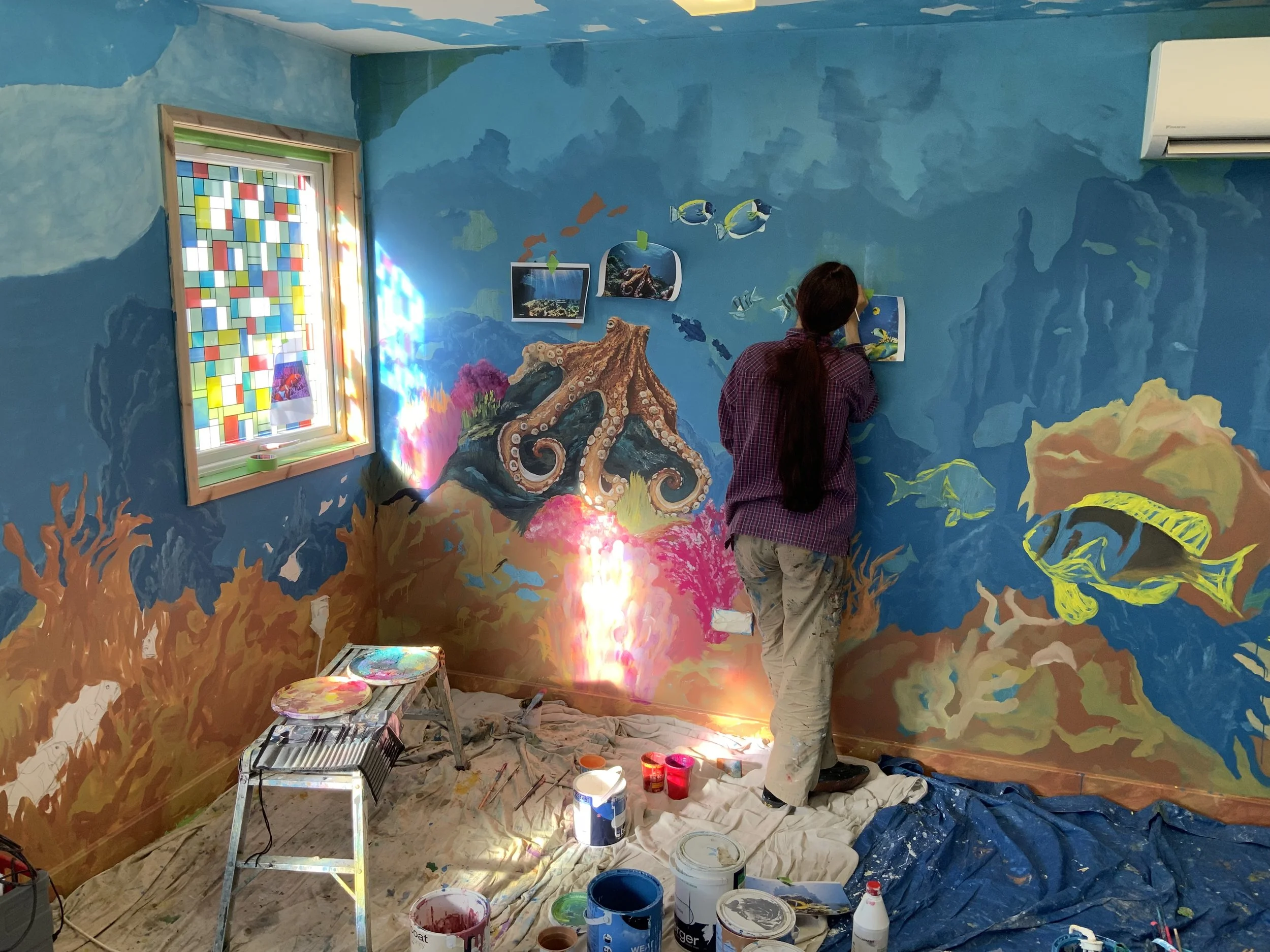 A person painting an underwater ocean mural on a blue wall with various sea creatures and coral scenery, with painting supplies on the floor and a stained glass window reflecting sunlight.