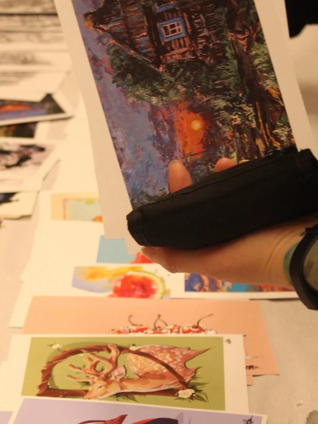 Person holding a colorful landscape painting in front of various other artworks hanging on a wall.