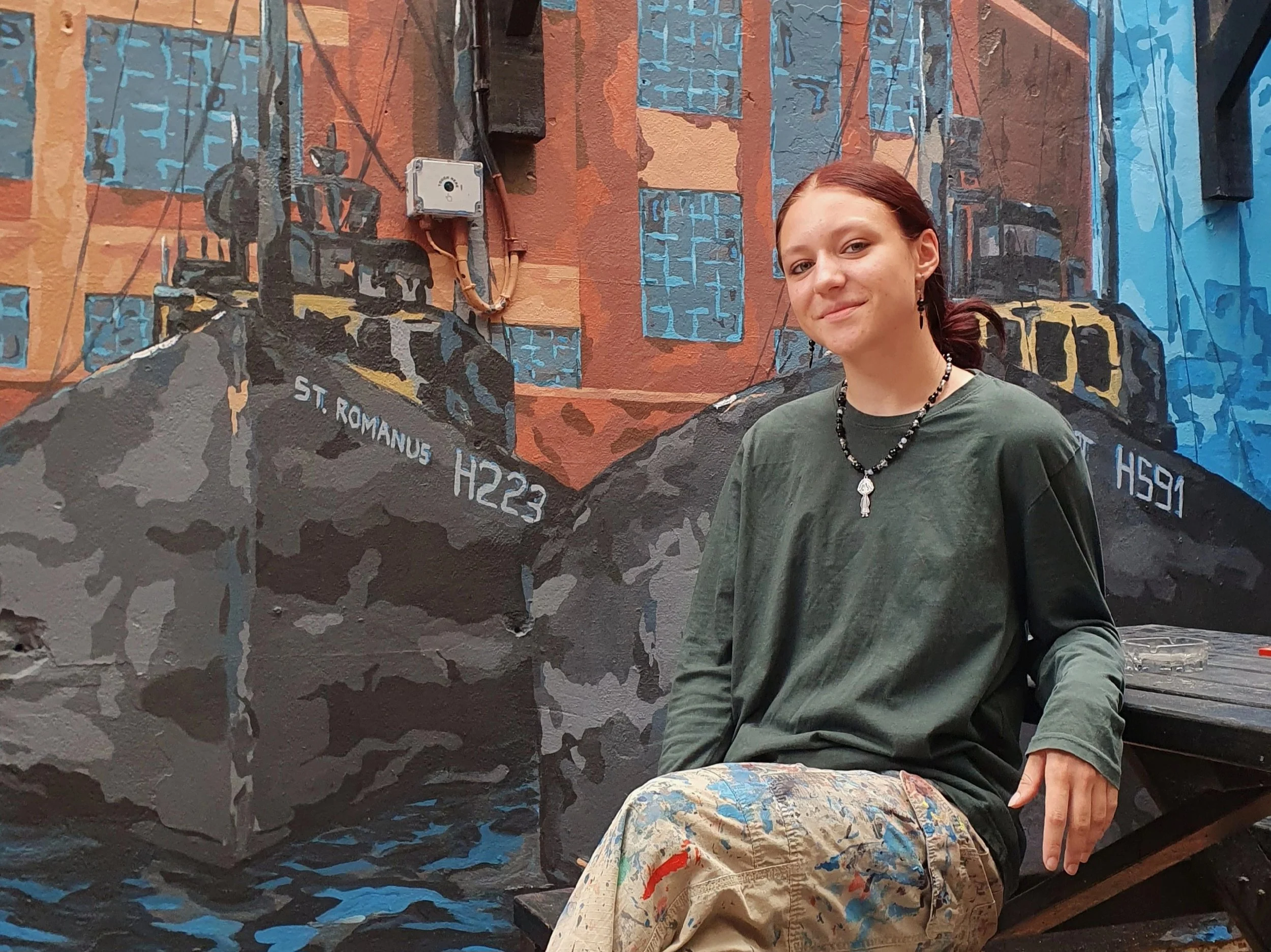 Young woman sitting in front of a colorful mural painted on a wall, wearing a green sweatshirt, necklace, and earrings, with painter's smock stained with paint at her lap.