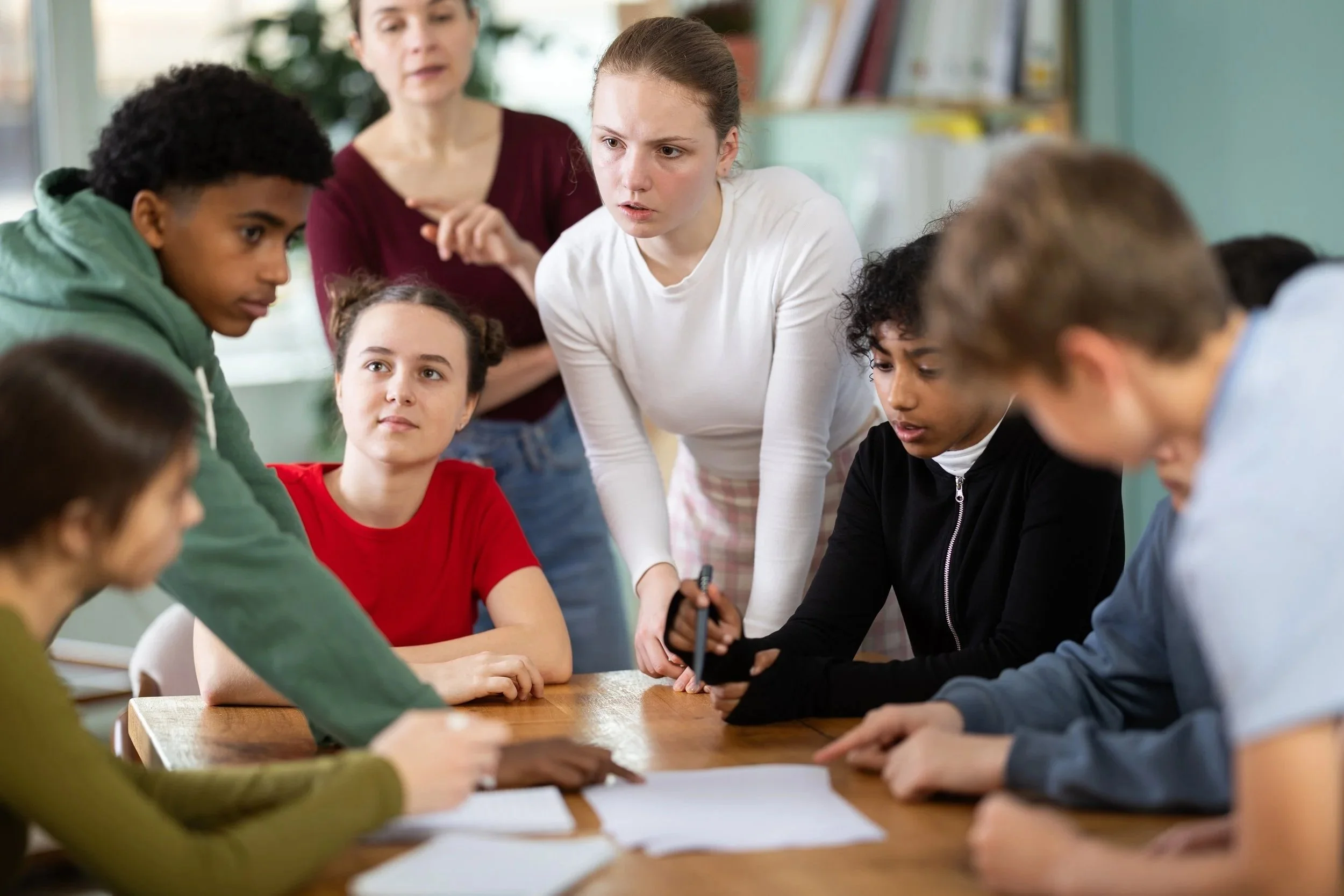 A group of diverse students and a teacher are gathered around a table in a classroom, engaged in a discussion or collaborative activity.